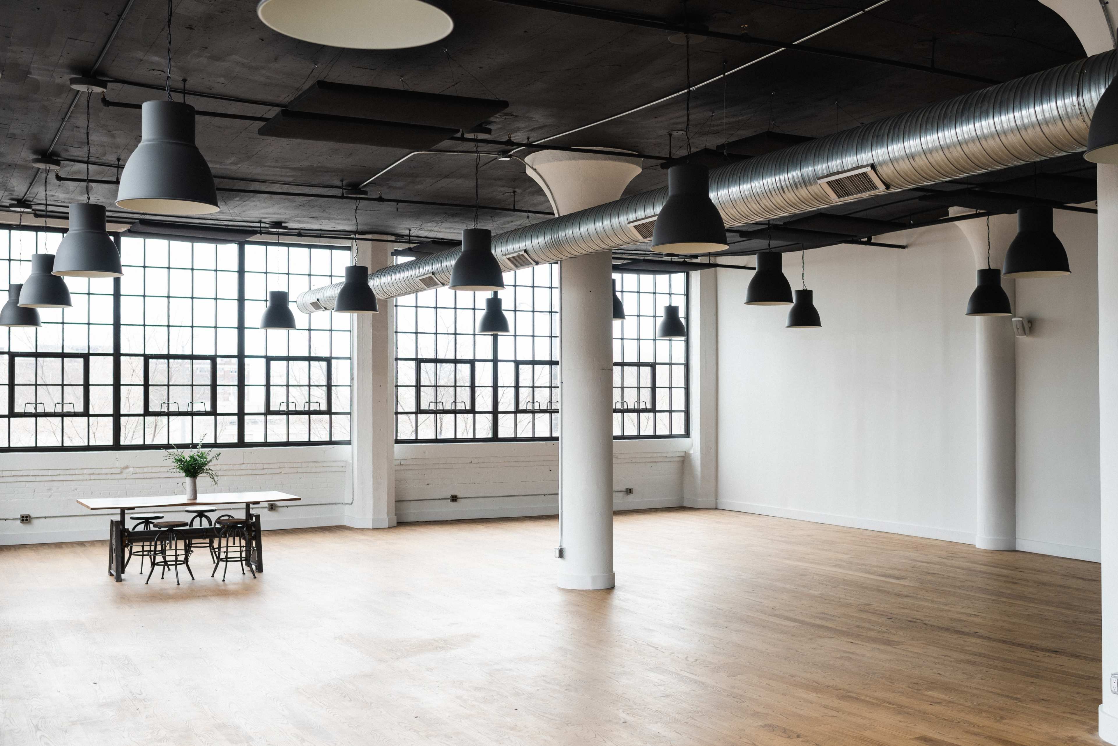A spacious, modern interior with large windows, exposed ductwork, and a wooden table surrounded by chairs.