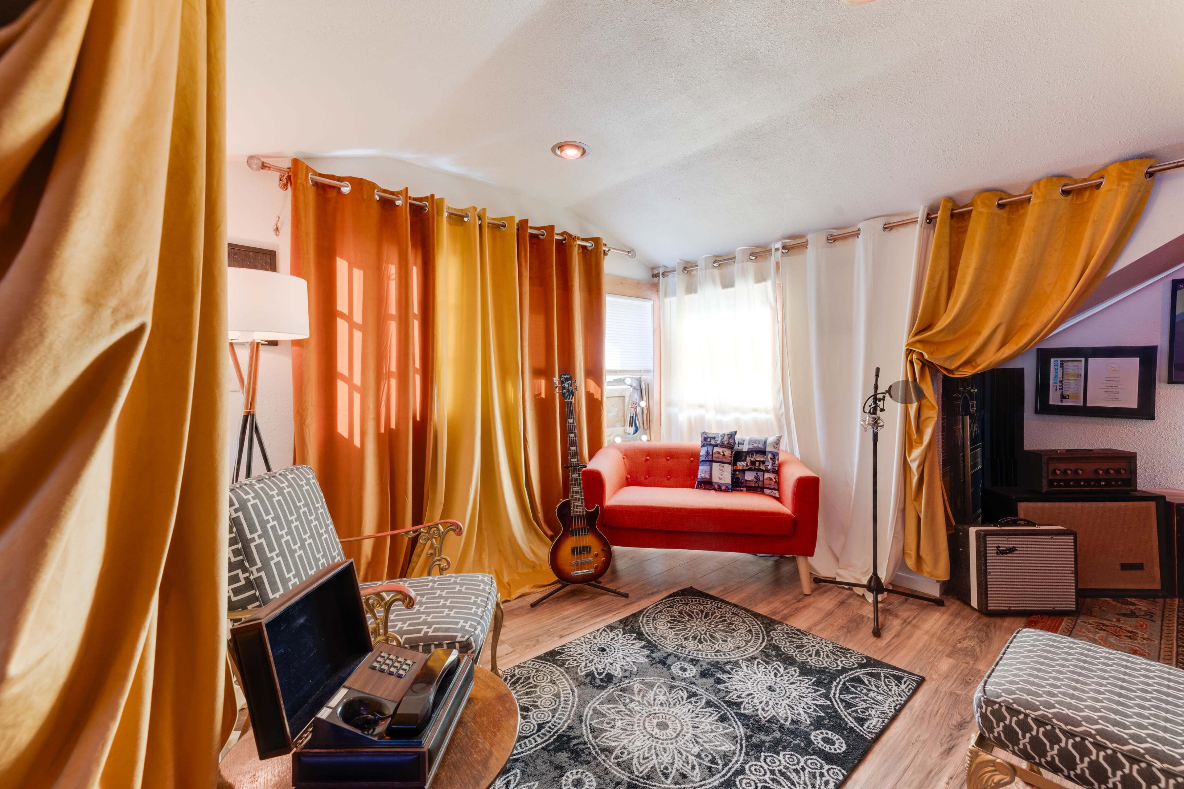 A cozy room features orange and yellow curtains, a red sofa, a patterned rug, and musical instruments.
