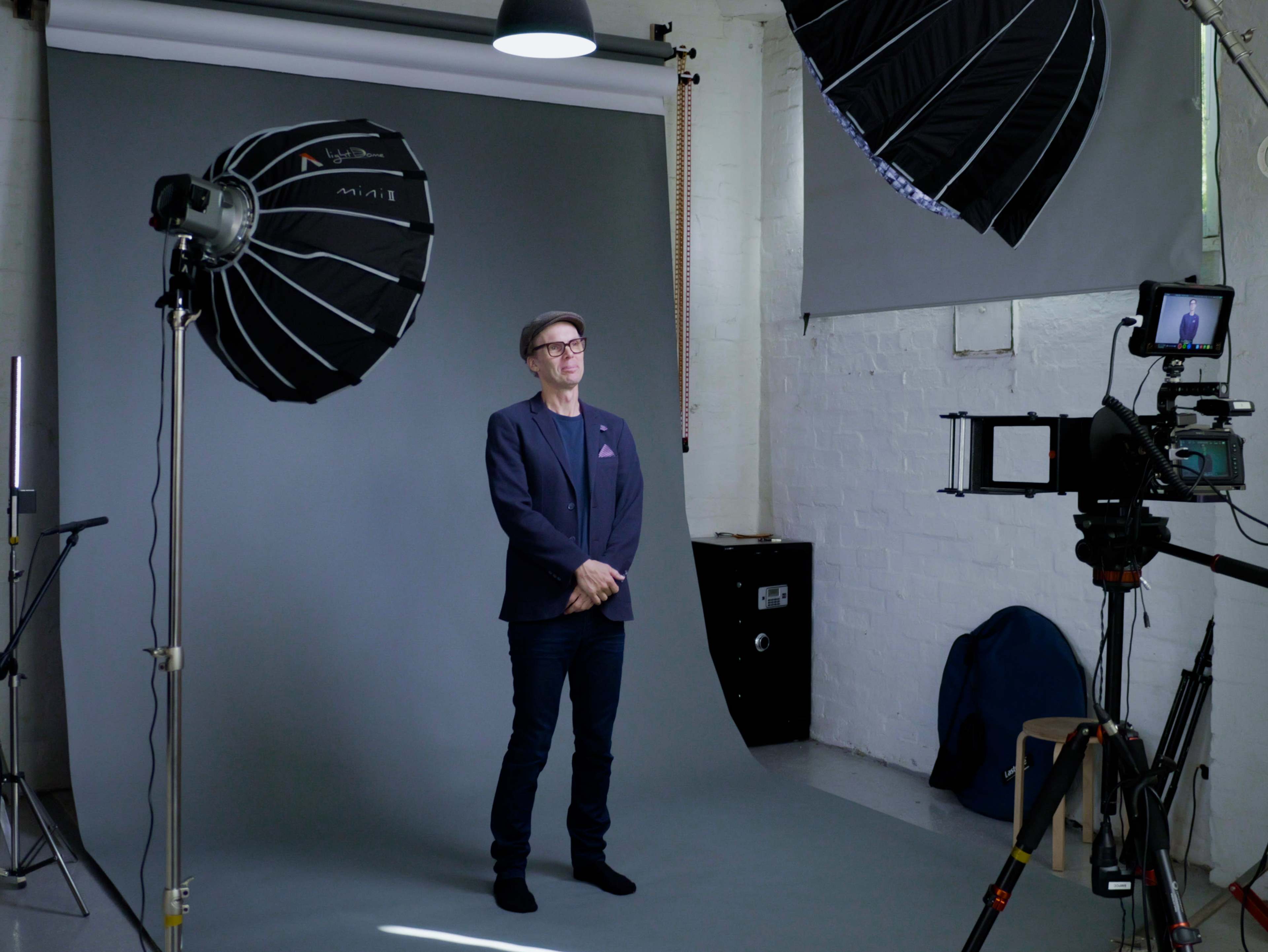 A man stands in a photography studio with a gray backdrop, flanked by large softbox lights and a camera on a tripod.
