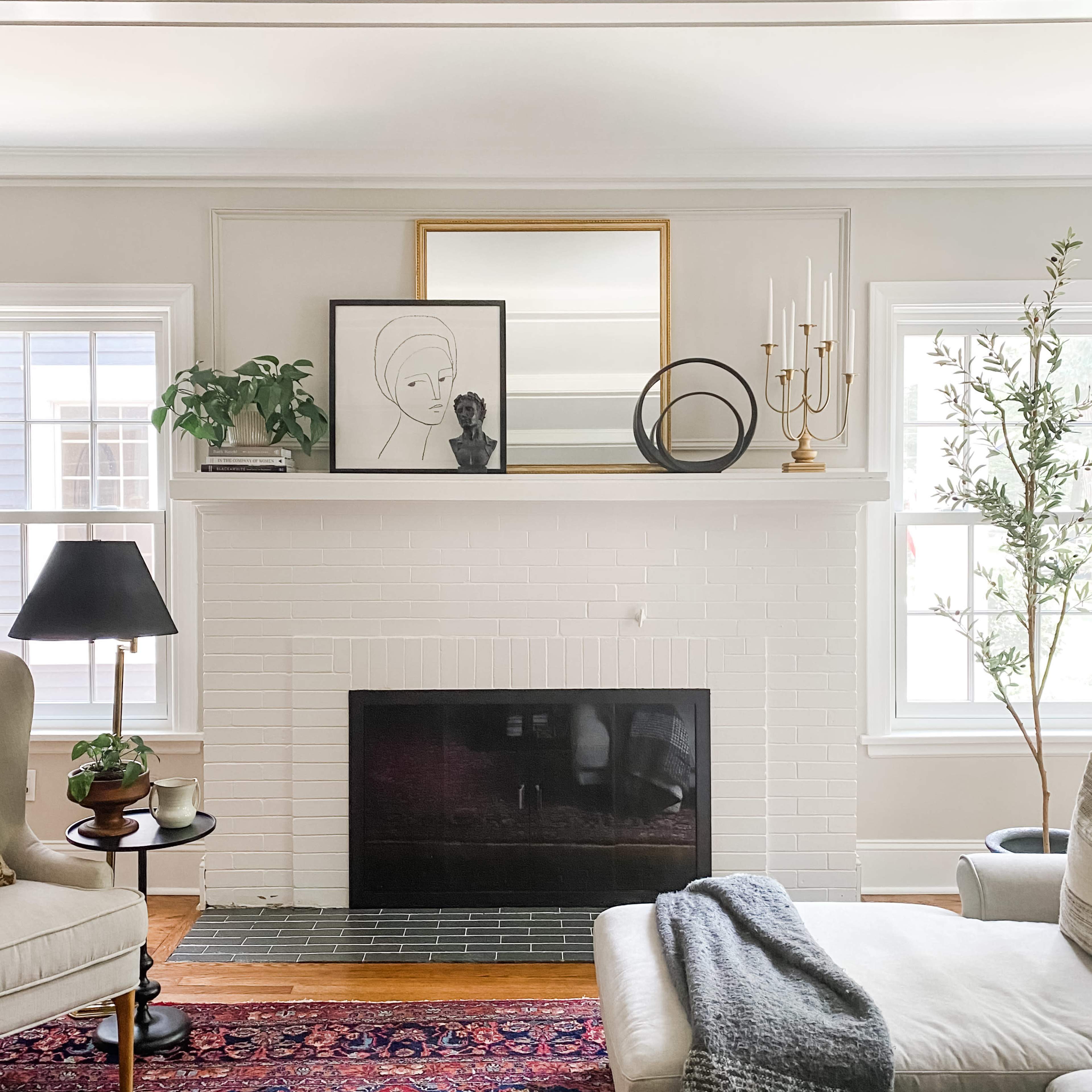 A modern living room with a white brick fireplace, framed artwork above it, a lamp on a side table, and a neatly arranged decorative plant beside large windows.