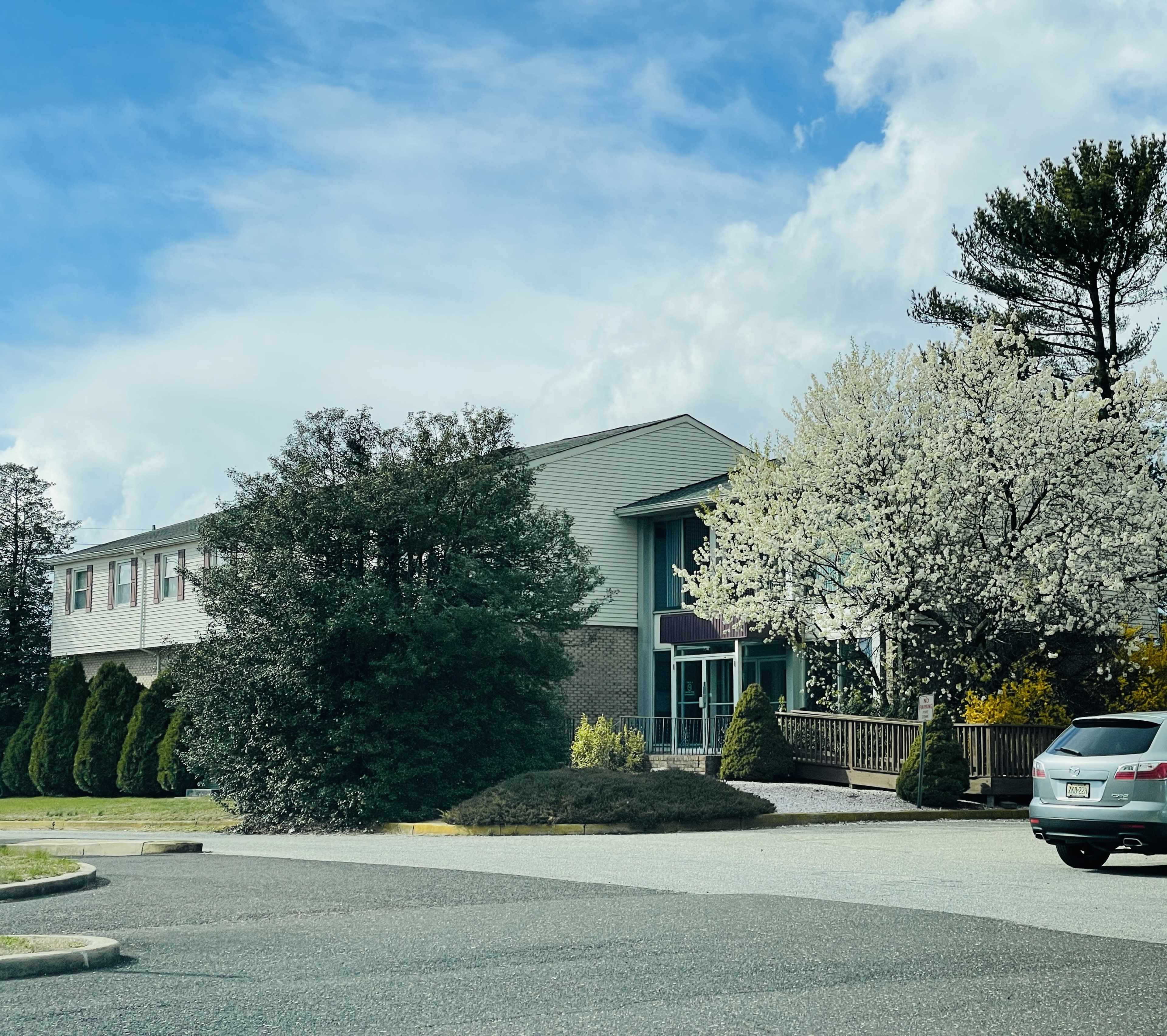 A building with a landscaped exterior featuring flowering trees and shrubbery is seen under a partly cloudy sky.
