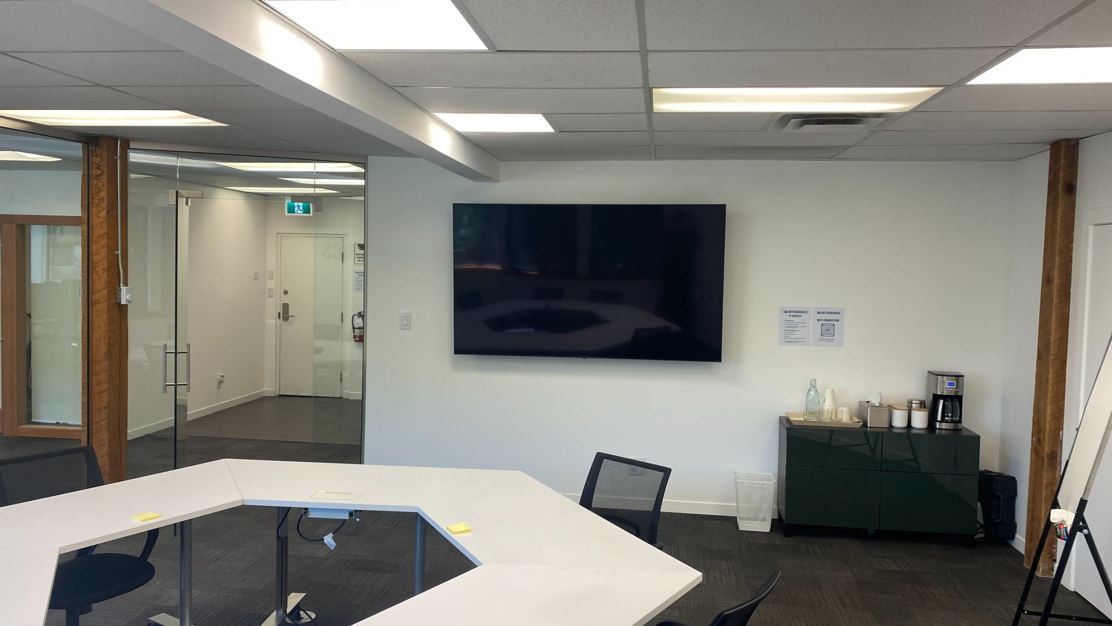 The image shows a modern office meeting room with a large wall-mounted television, a white hexagonal table, and a coffee station against a wall.