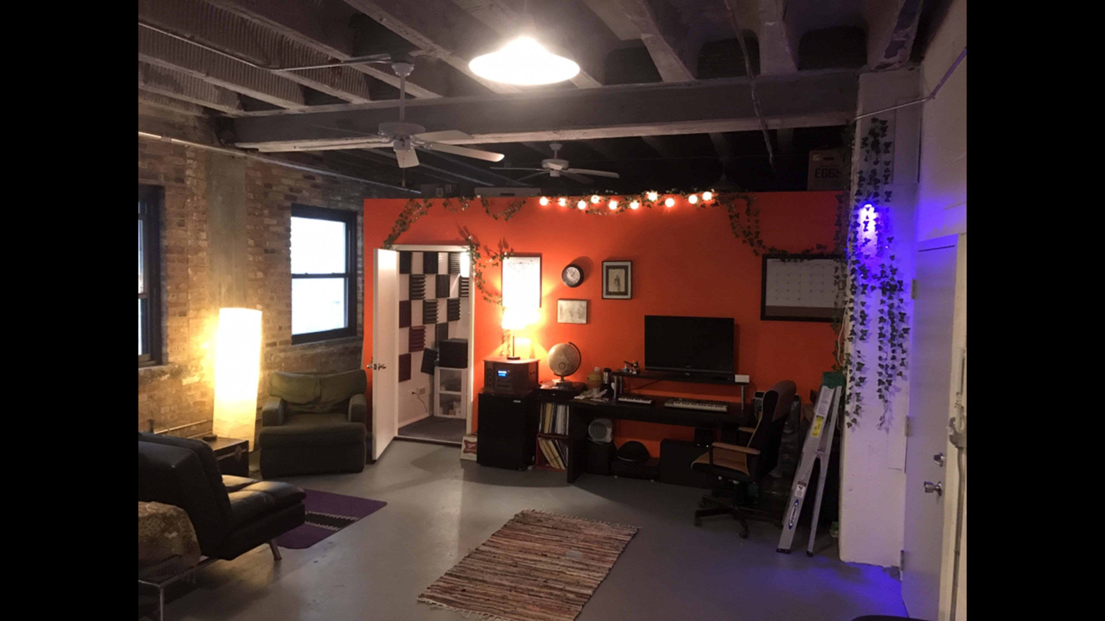 The image shows a cozy room with an orange accent wall, a desk with a computer, a green couch, and plants decorating the space alongside various light sources.