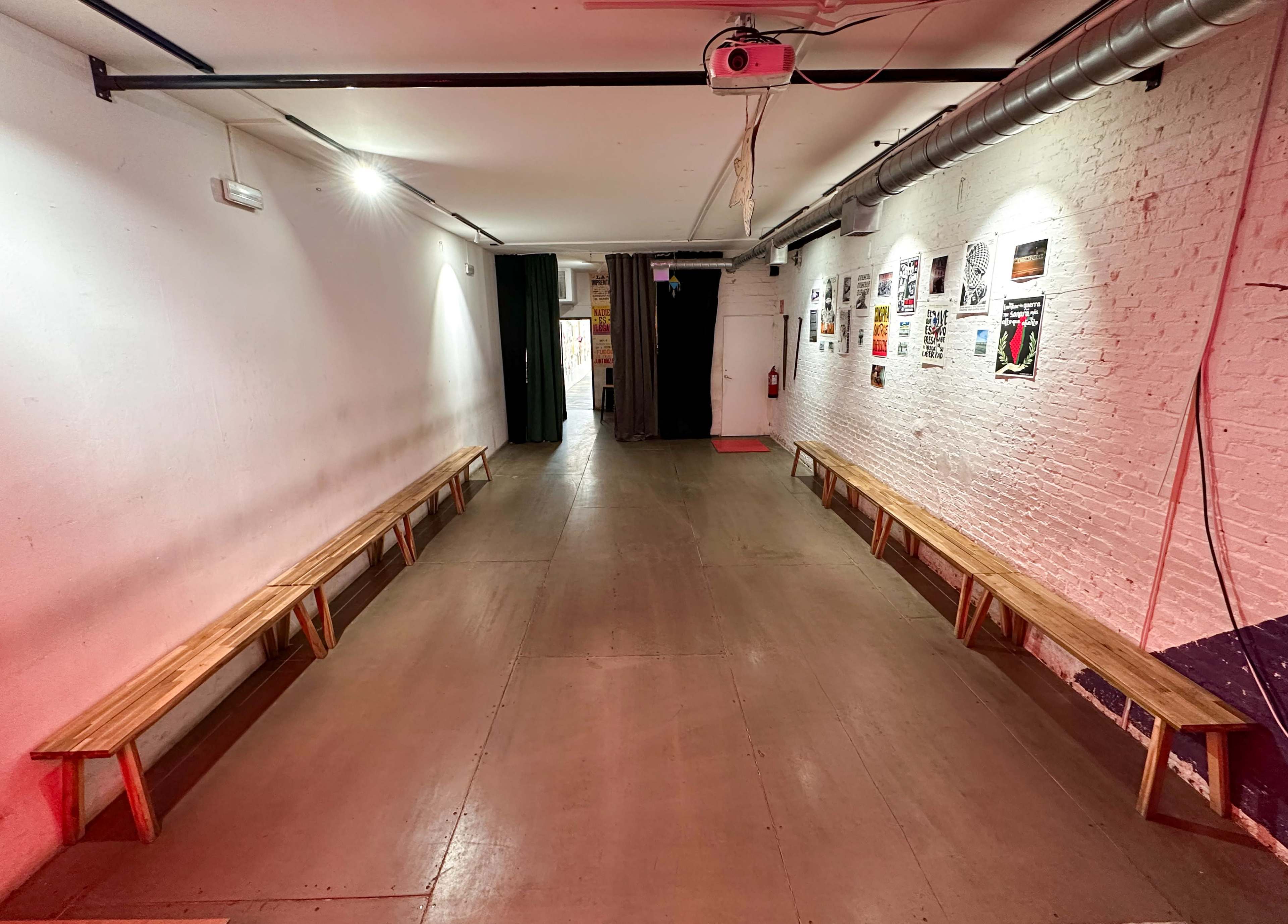 The image shows a long, narrow hallway with wooden benches on either side and a series of posters on the wall, illuminated by overhead lights.