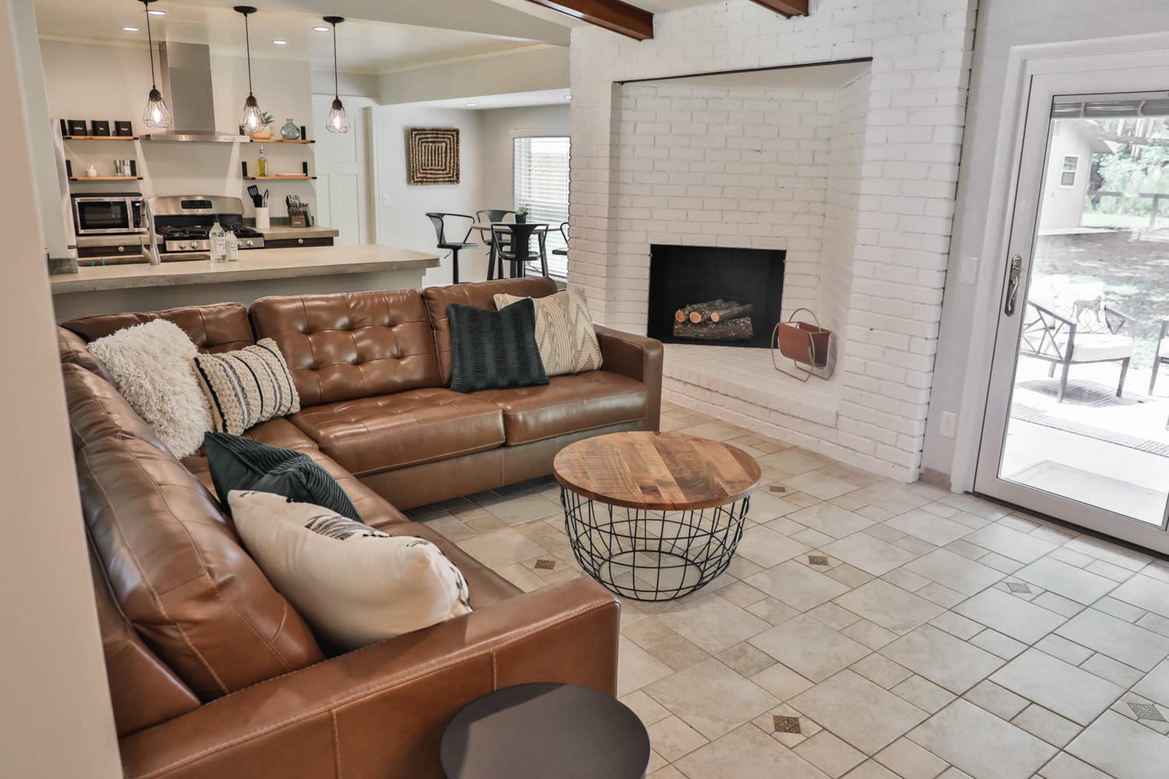 A cozy living room featuring a brown leather sectional sofa, a circular wooden coffee table, and a white brick fireplace.