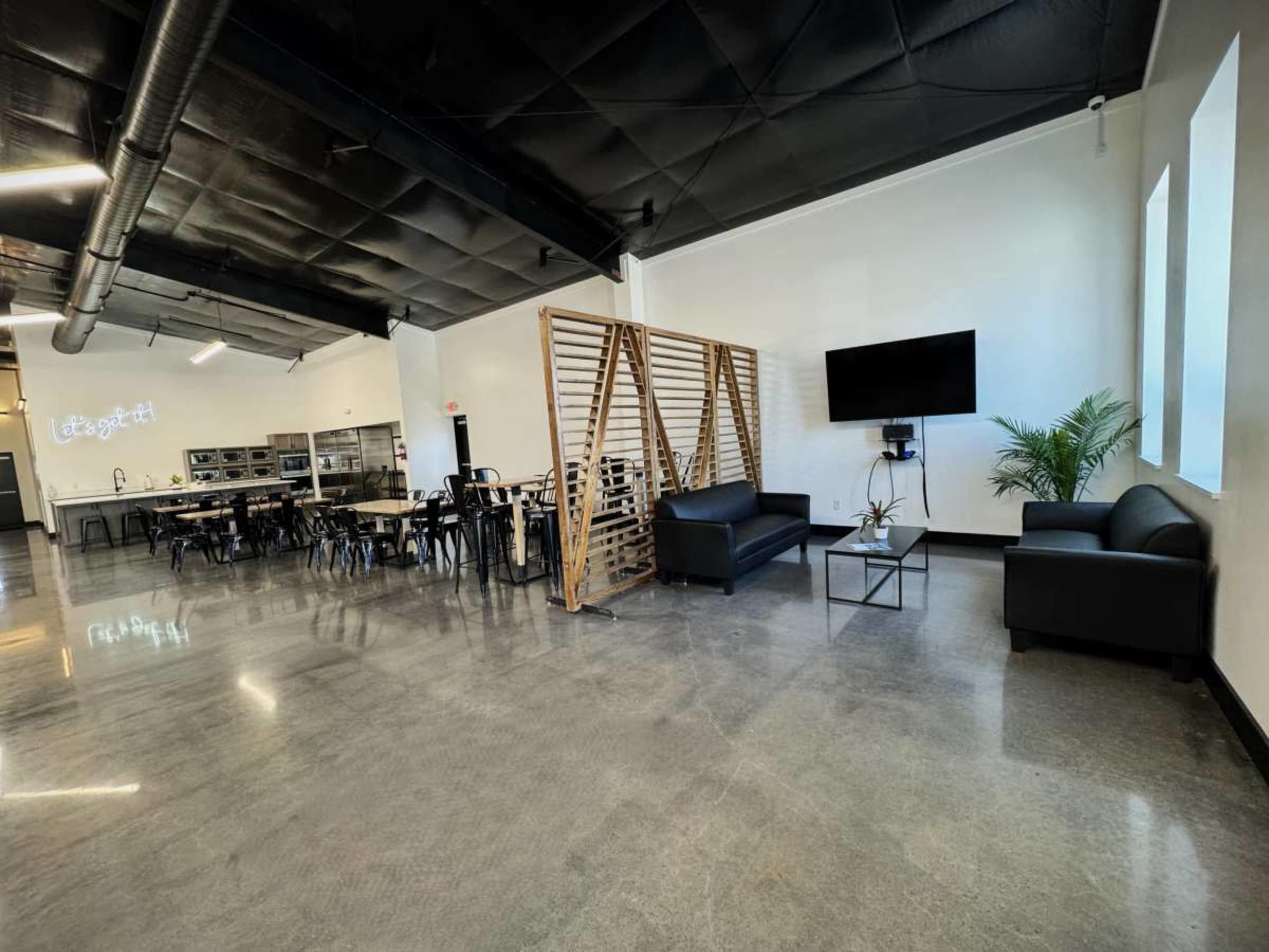 A modern indoor space featuring a seating area with two black sofas, a television mounted on the wall, and a dining area with tables and chairs in the background.