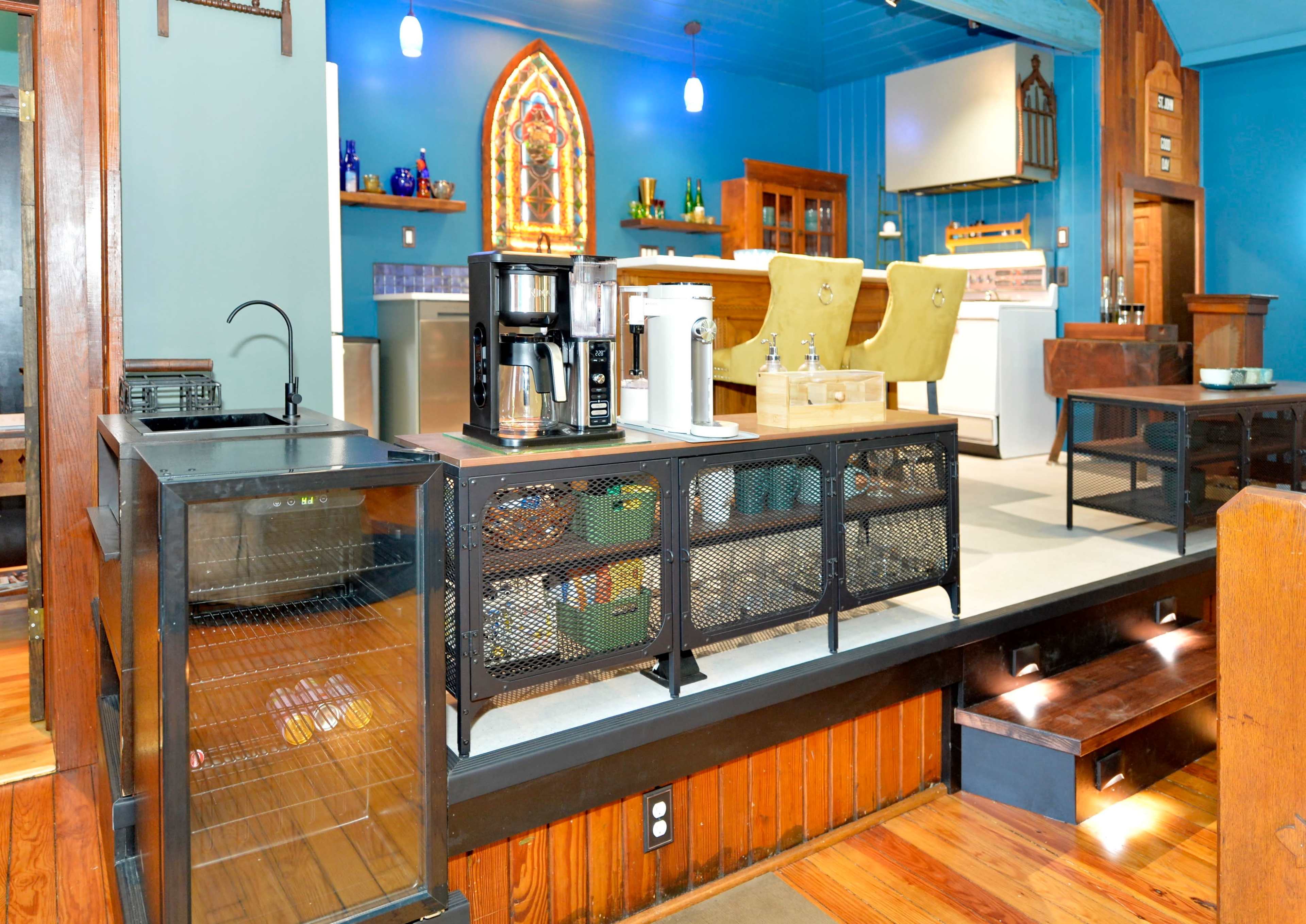 The image shows a brightly colored café interior featuring a coffee station and seating area, with wooden floors and decorative elements on the walls.