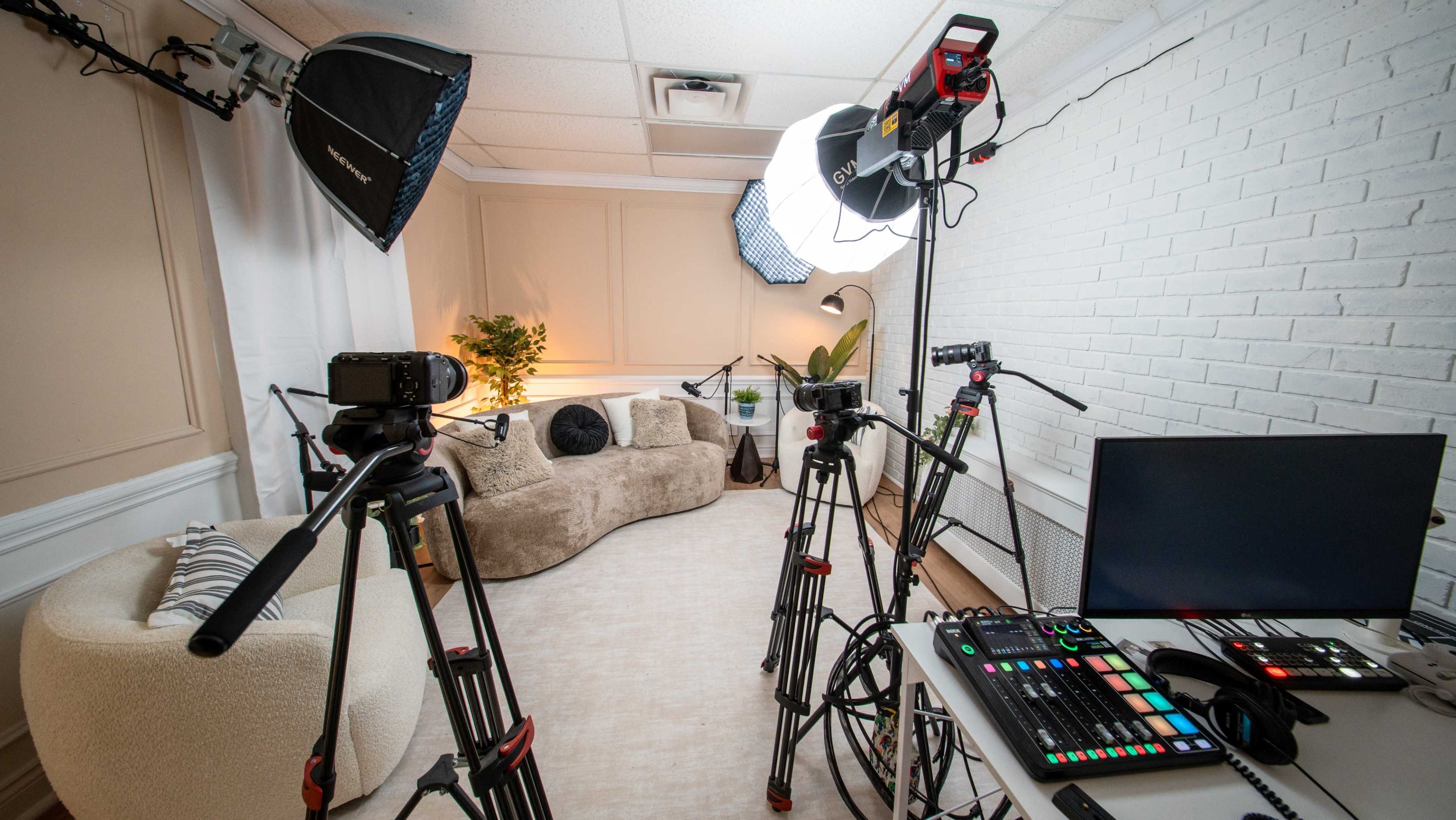 The image shows a well-equipped video production studio featuring multiple cameras on tripods, studio lights, and a computer setup on a table, with a cozy seating area in the background.