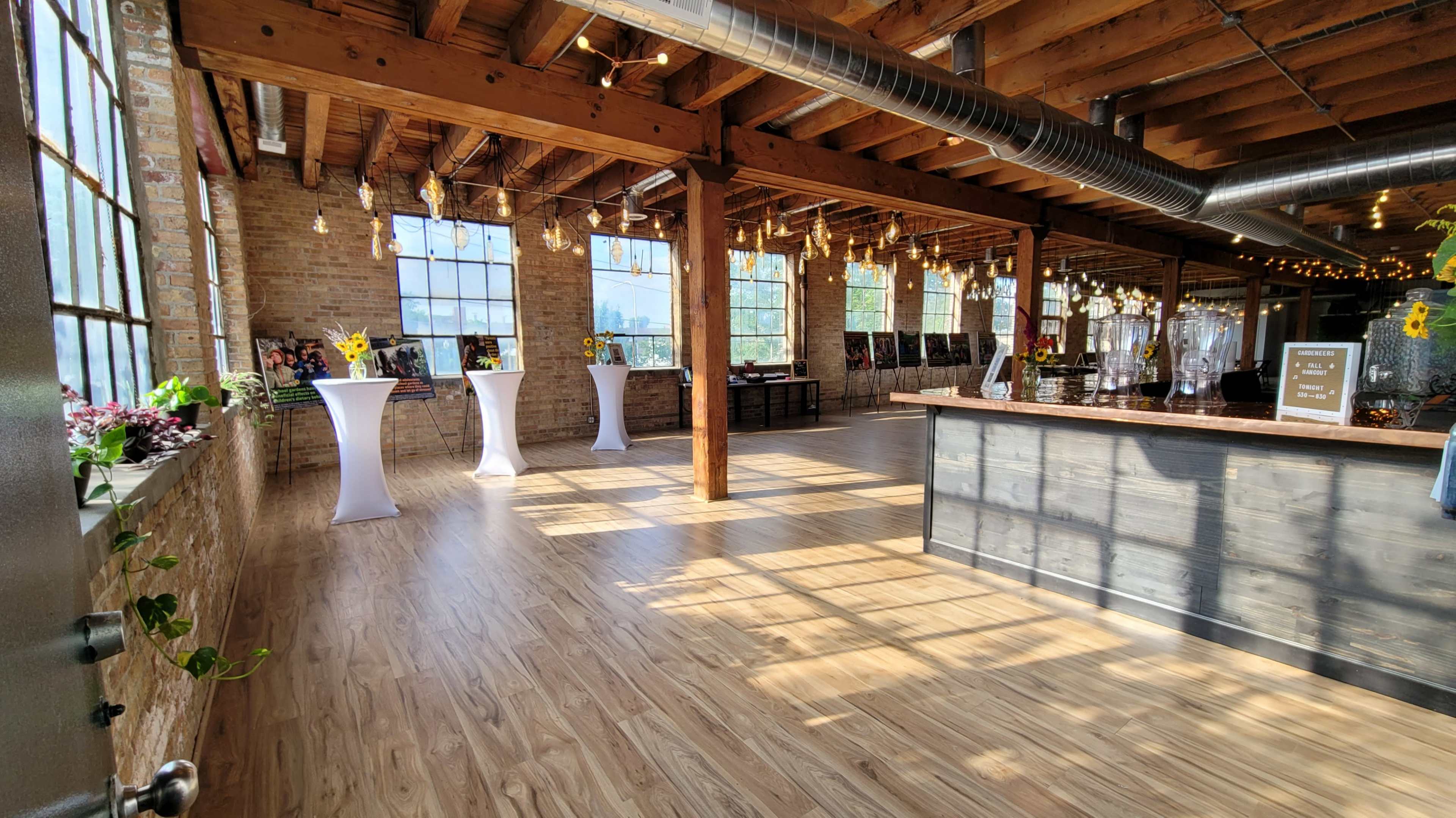 The image shows a spacious room with wooden beams, large windows, and minimalistic furniture, featuring light streaming in and decorative elements like sunflowers.