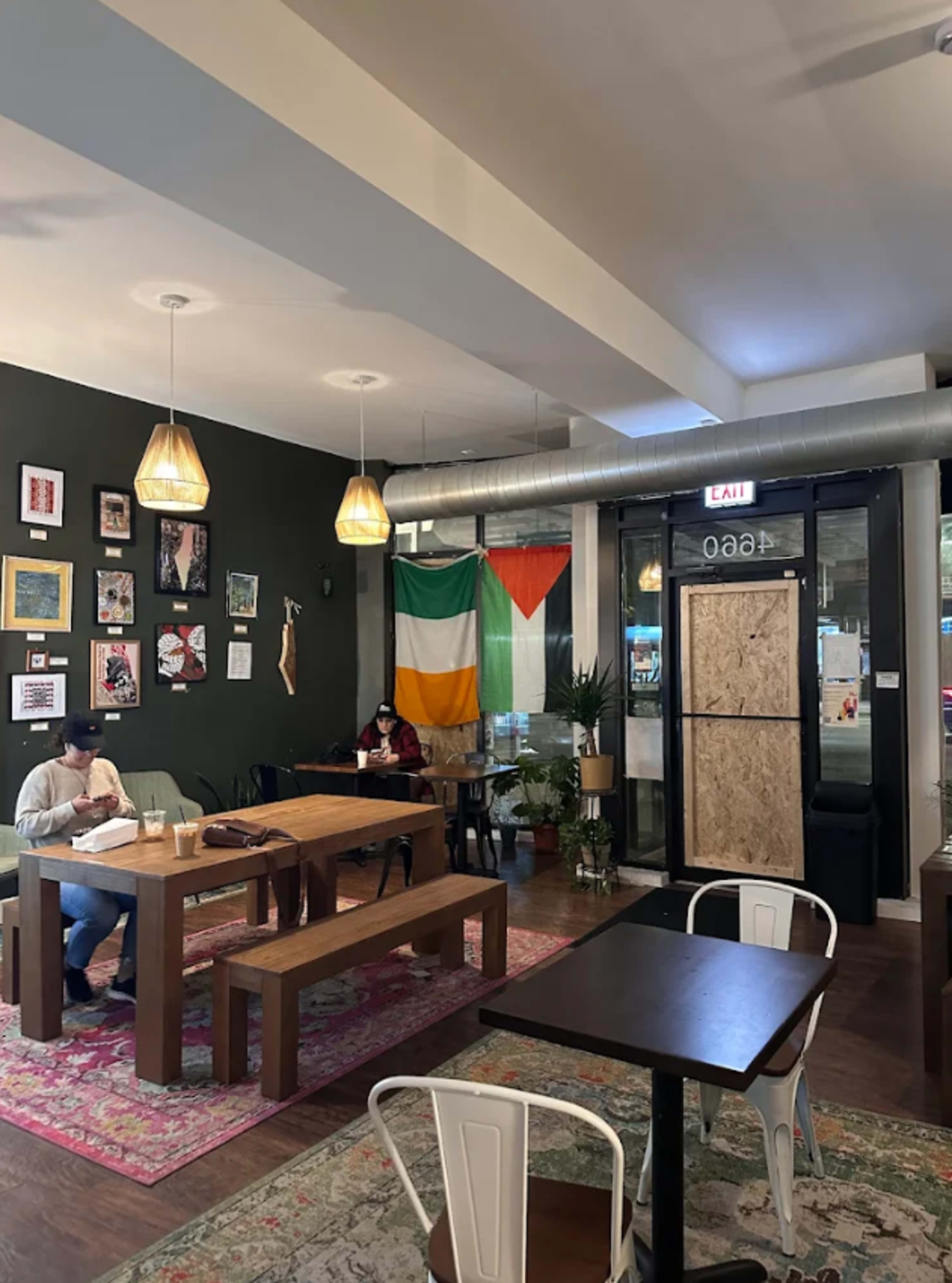 The image shows a cozy cafe interior with wooden tables, a large green and orange flag on the wall, and patrons seated, one writing and another using a laptop.
