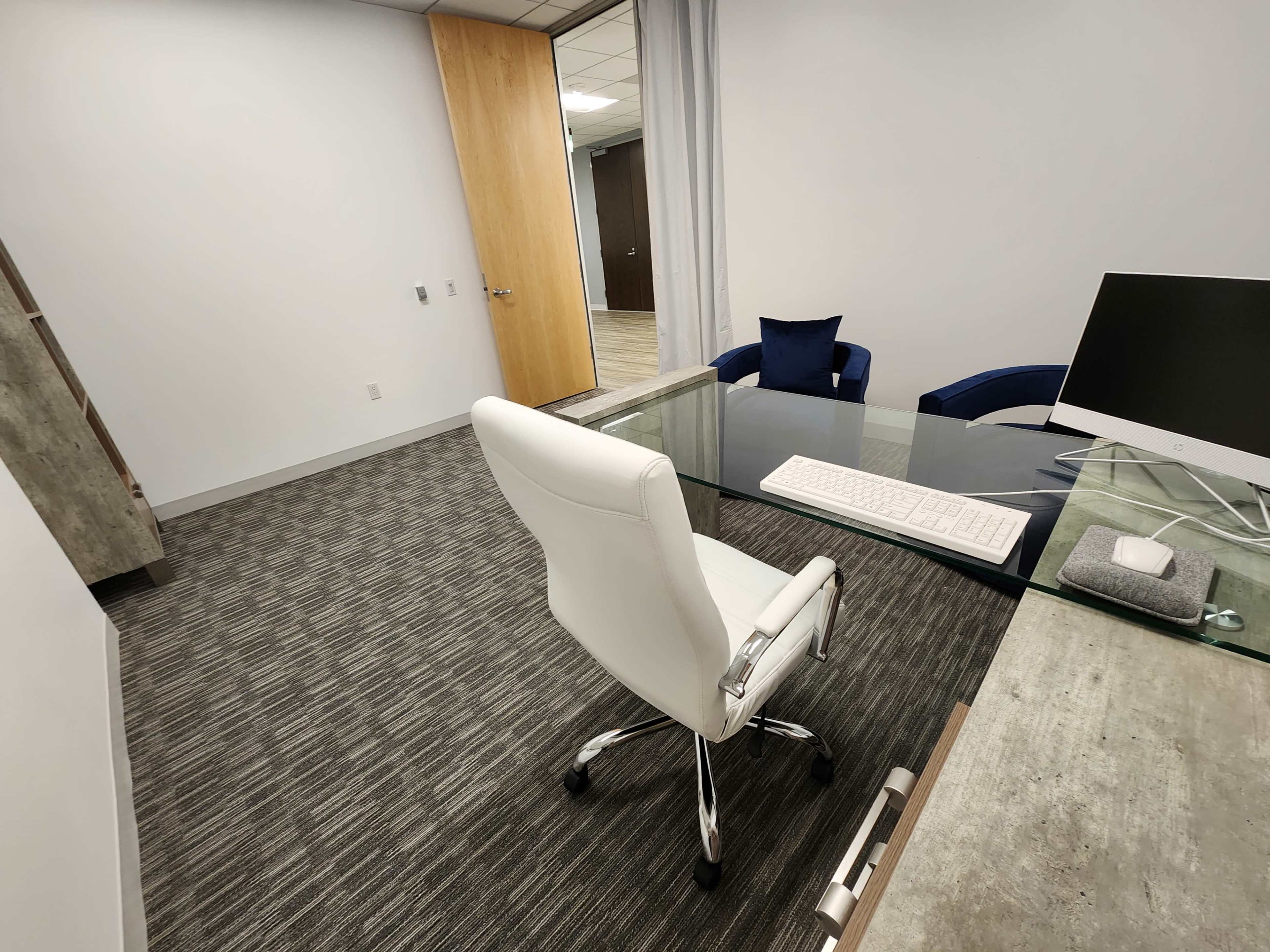 The image shows a modern office setting featuring a white ergonomic chair, a glass desk with a keyboard, and a computer monitor, alongside a plush blue chair and a wooden door.
