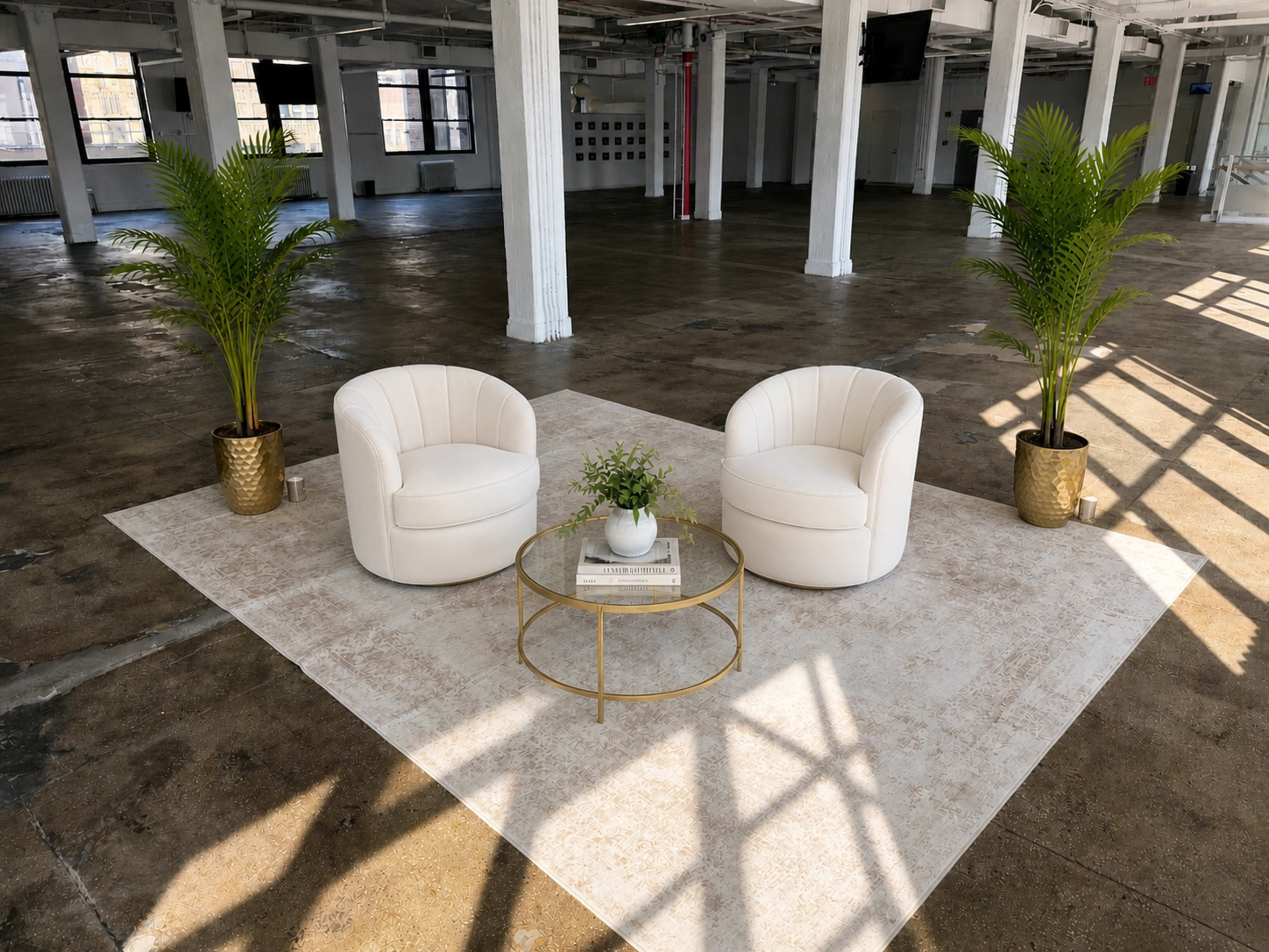 The image shows two white, upholstered chairs facing each other on a light rug next to a small round table, all situated in an empty, well-lit industrial space with large windows.