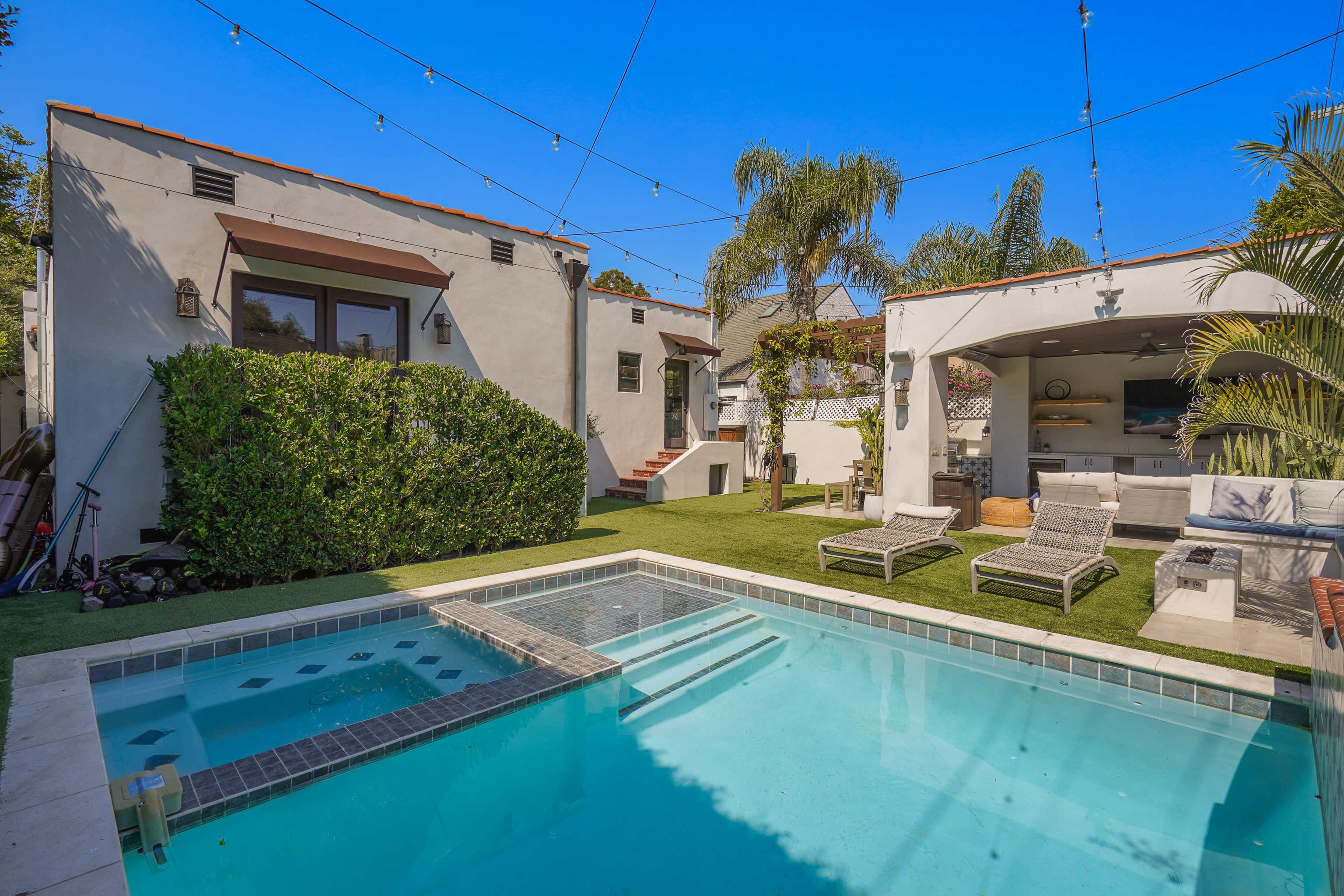 The image shows a backyard featuring a swimming pool, outdoor seating areas, and surrounding greenery with a clear blue sky overhead.