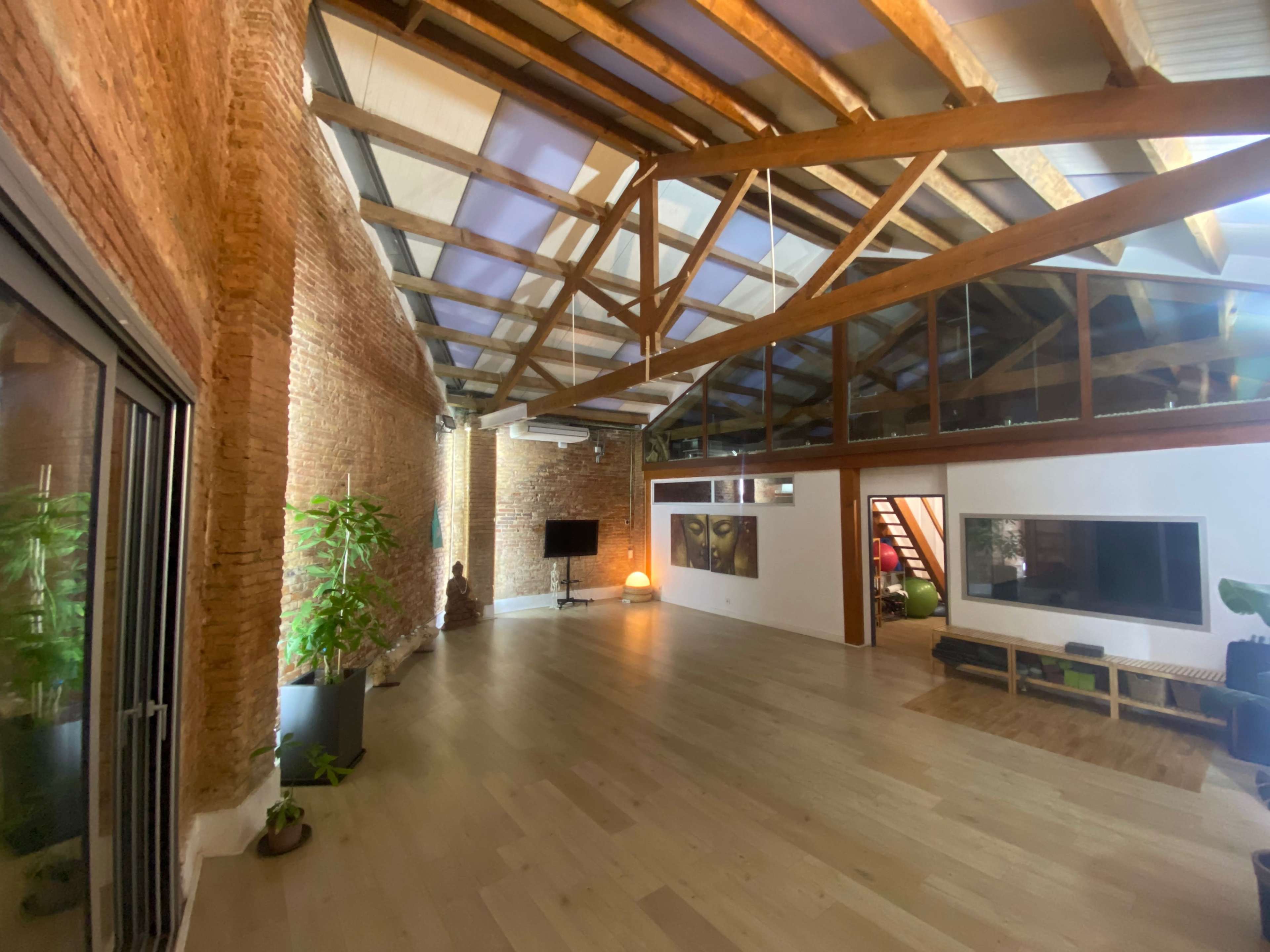 The image shows a spacious loft with exposed brick walls, wooden beams, and large windows, featuring a minimalist design and a few plants.