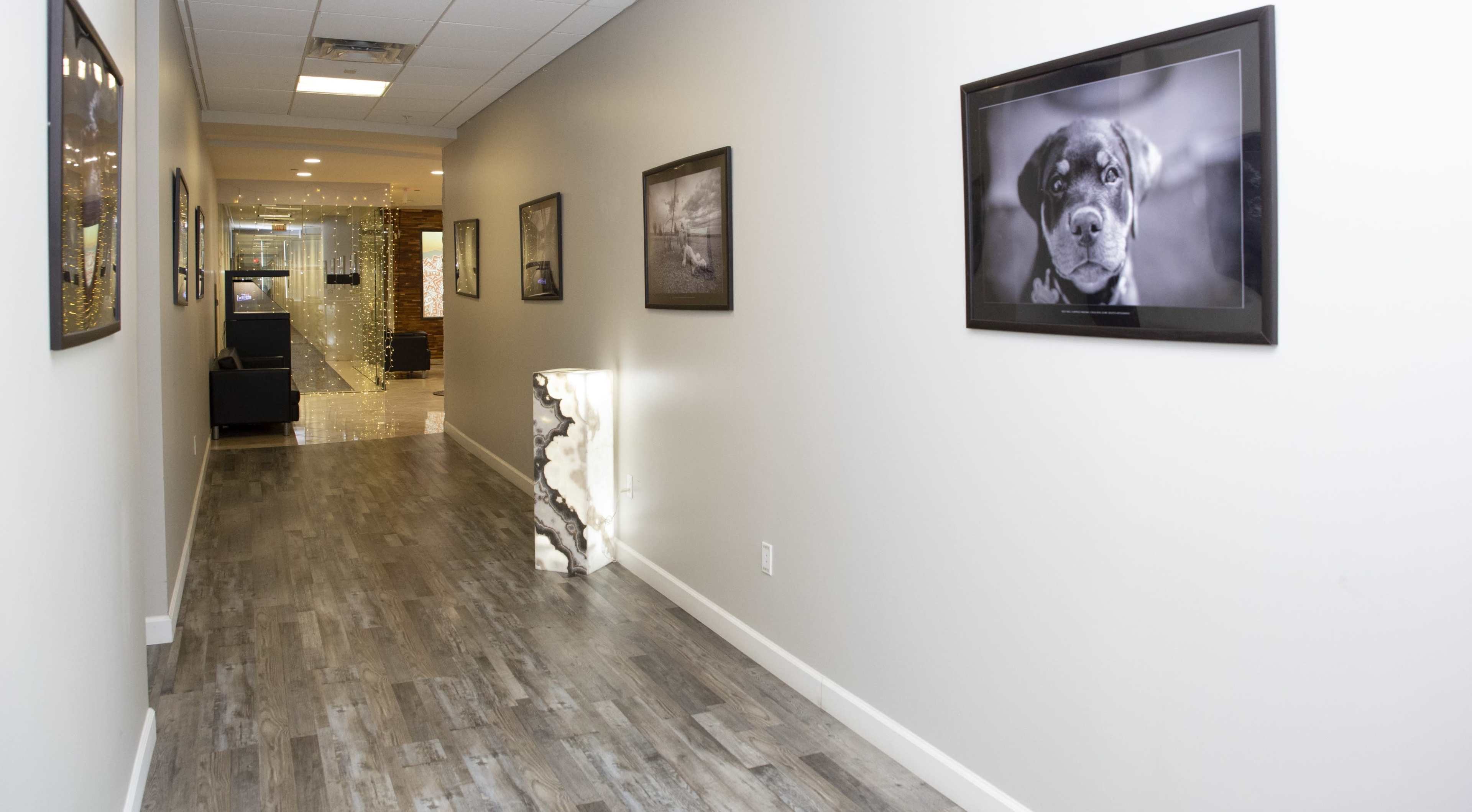 A well-lit hallway with wooden flooring, lined with framed black-and-white photographs, and a modern lamp at the end.