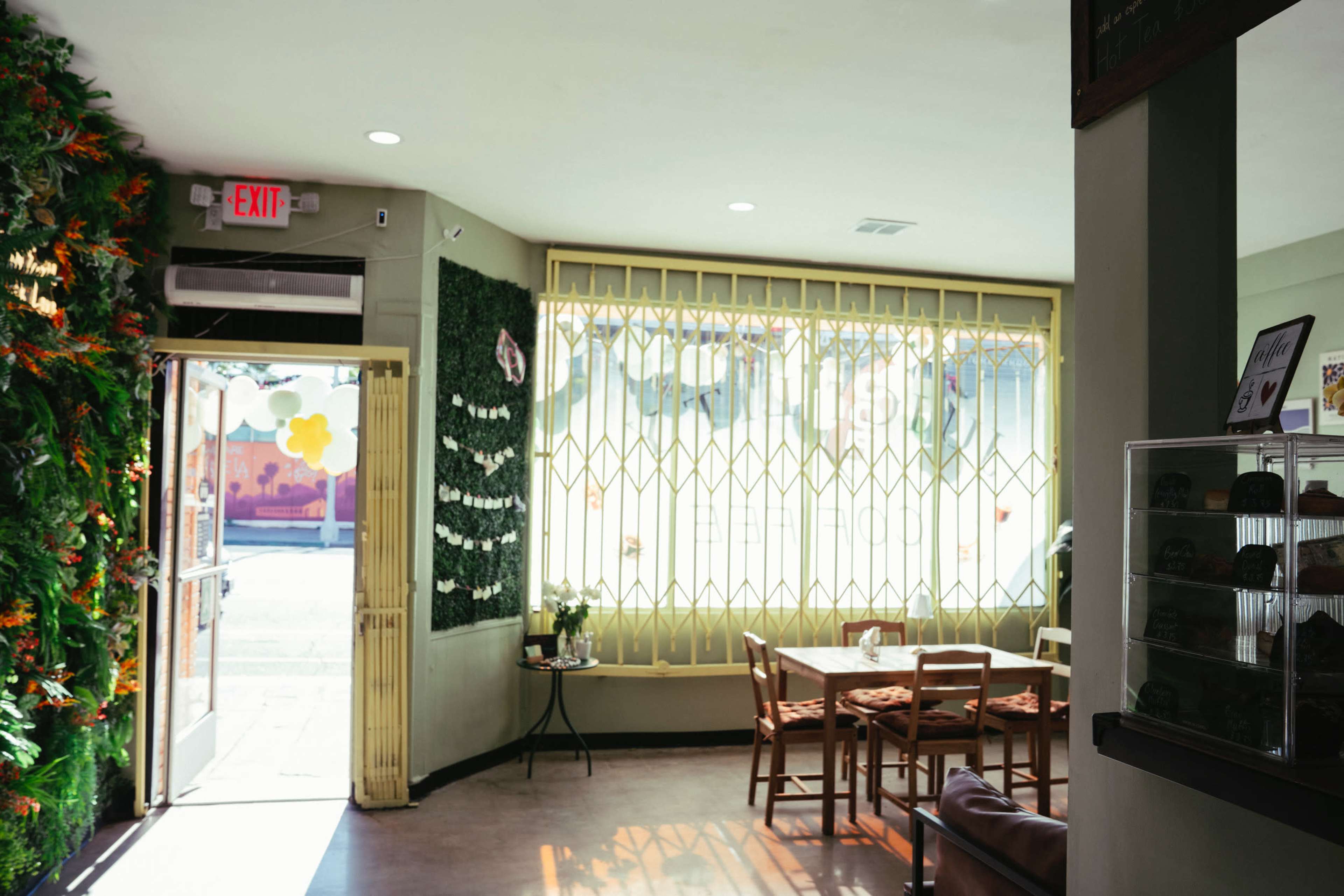 The image shows the interior of a cafe featuring a green wall with plants, a table and chairs, and a large, gated window allowing natural light inside.