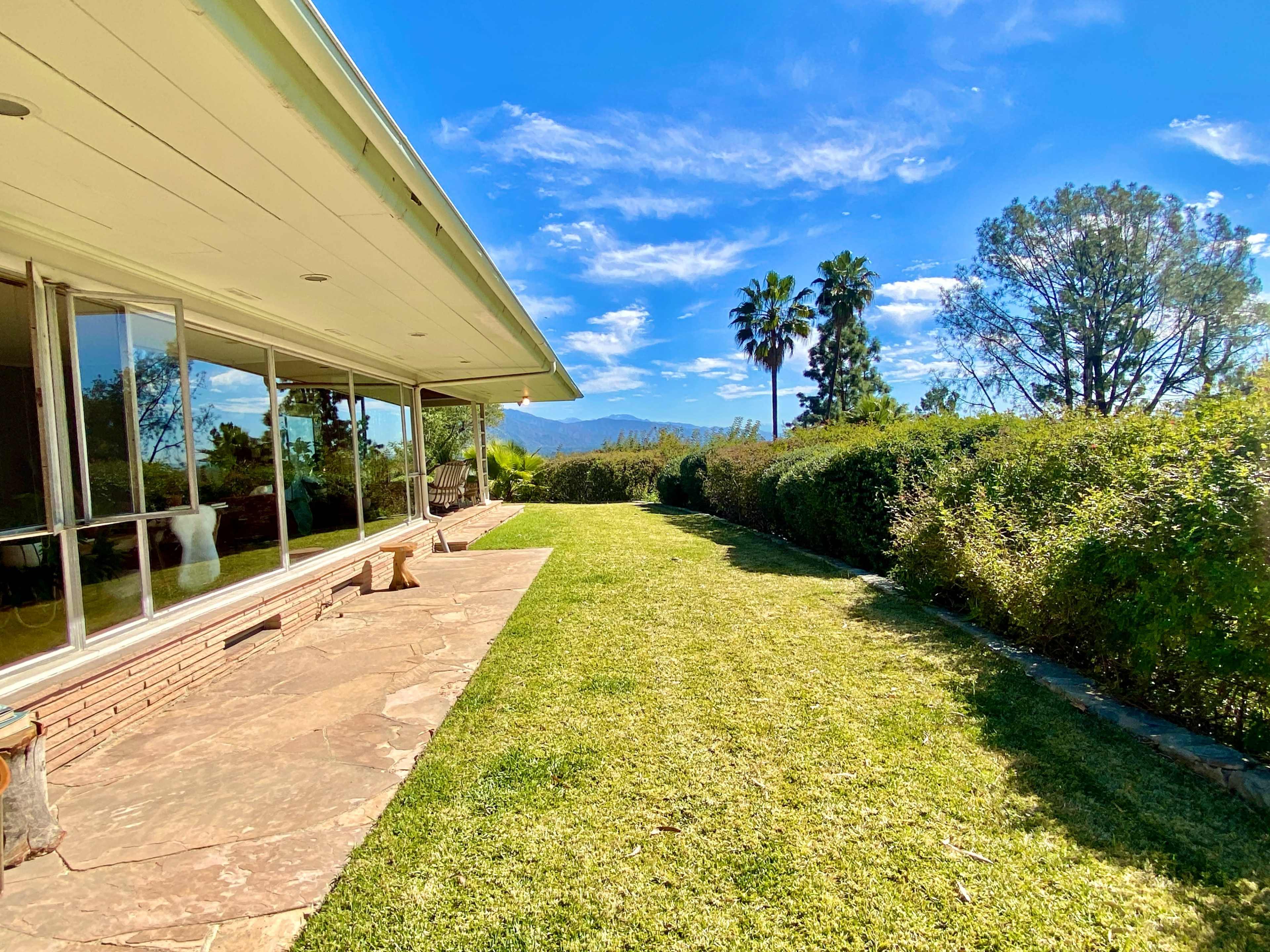 The image shows a landscaped yard with a grassy area and a patio adjacent to a modern house, framed by trees and distant mountains under a clear sky.