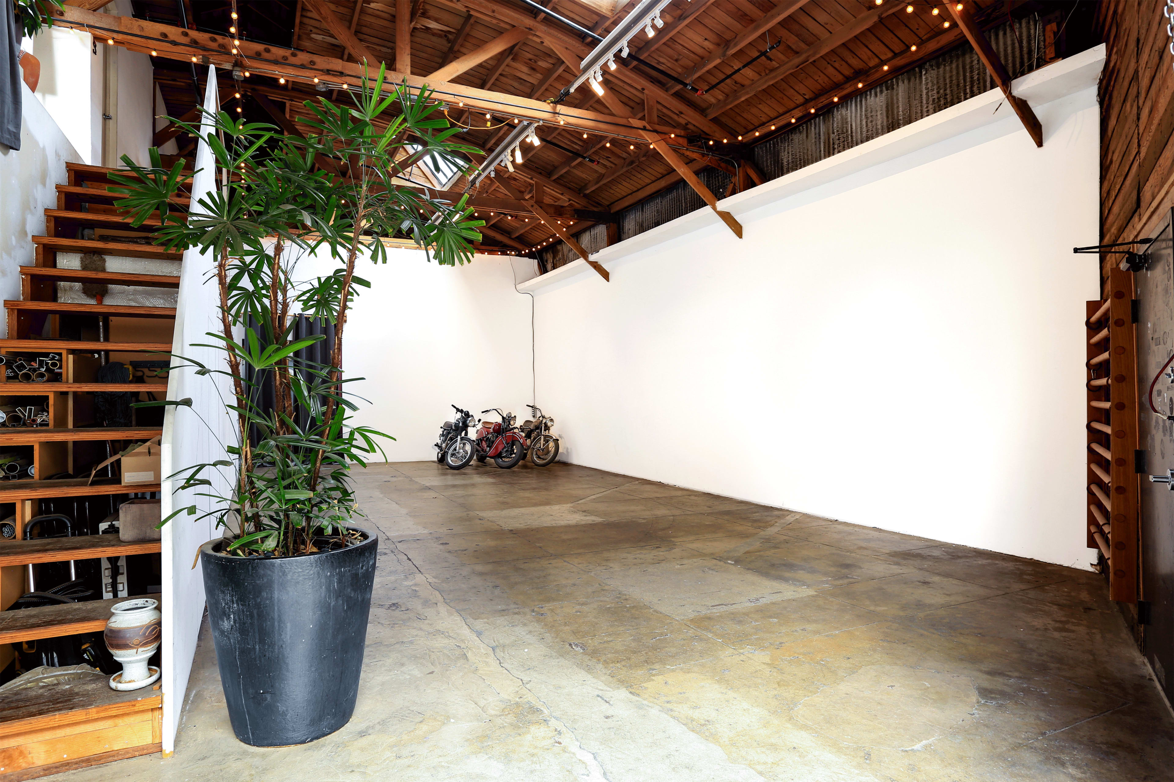The image shows a spacious, well-lit interior with a staircase on the left, a large potted plant, and three motorcycles parked against a white wall.