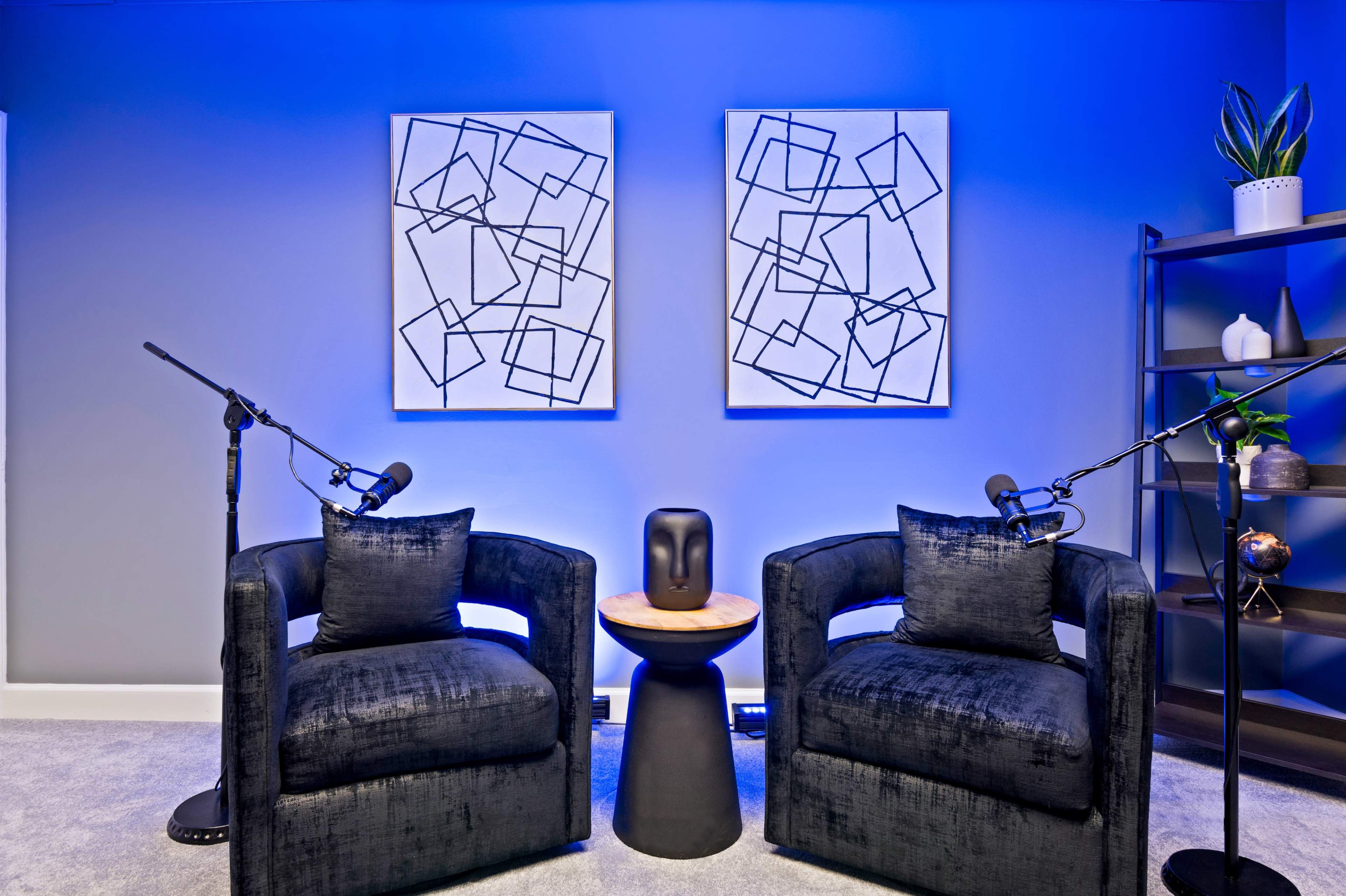 Two black armchairs positioned facing a small round table, with abstract artwork on the walls and microphones set up for recording.