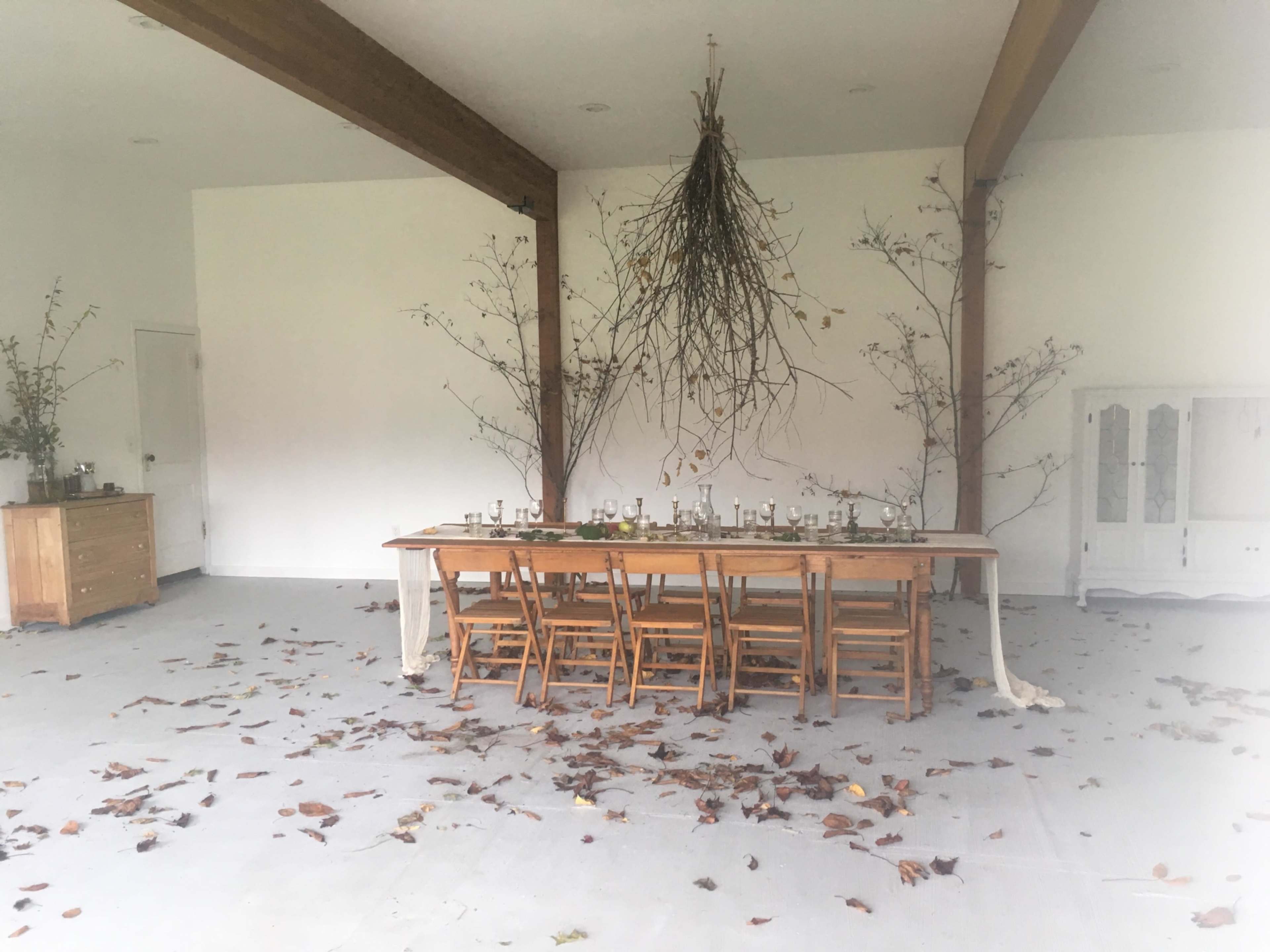 A long wooden table with chairs is set in a room decorated with autumn leaves and branches hanging from the ceiling.