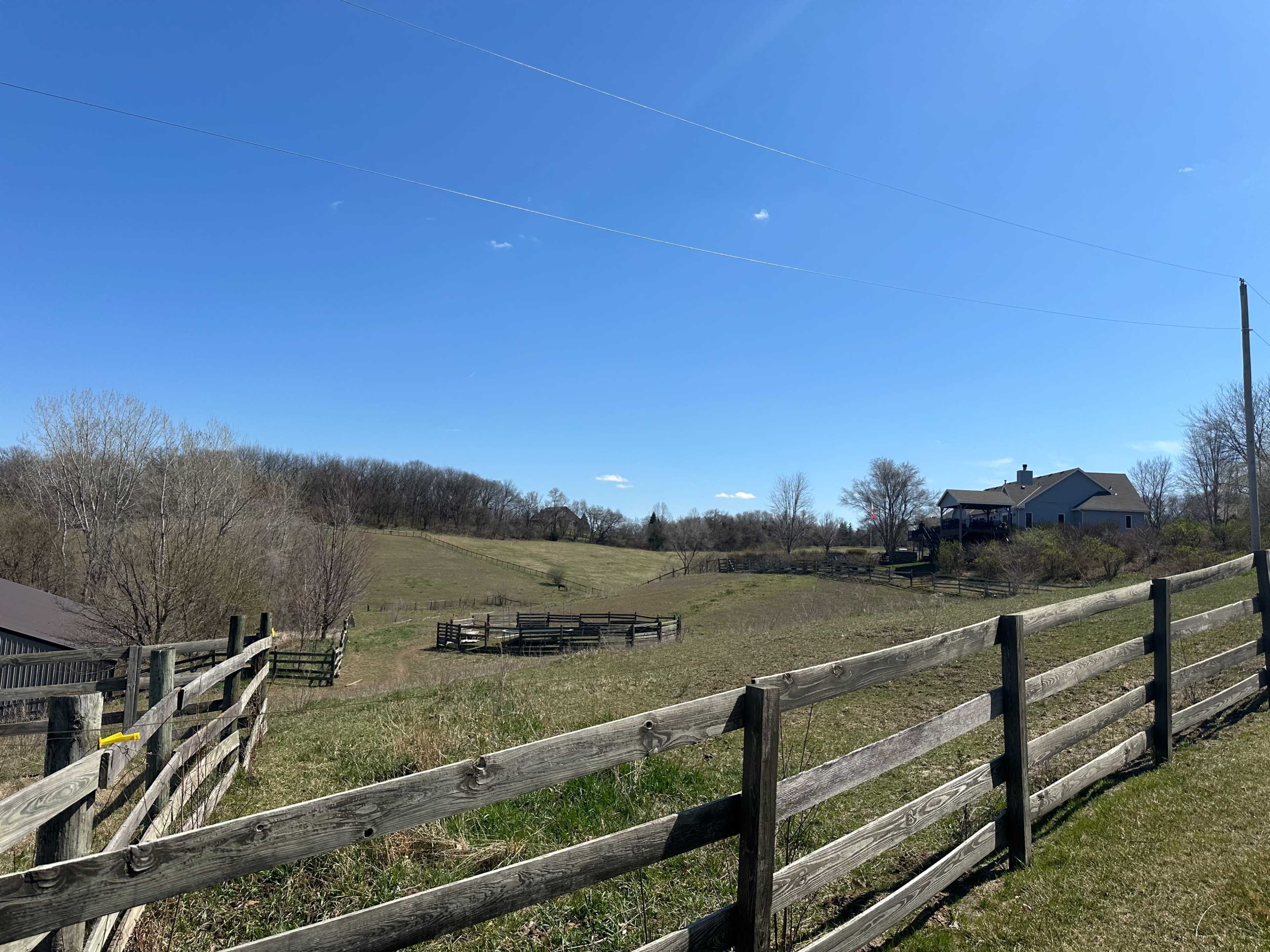 A rural landscape features a grassy field with a wooden fence, leading to homes and trees under a clear blue sky.