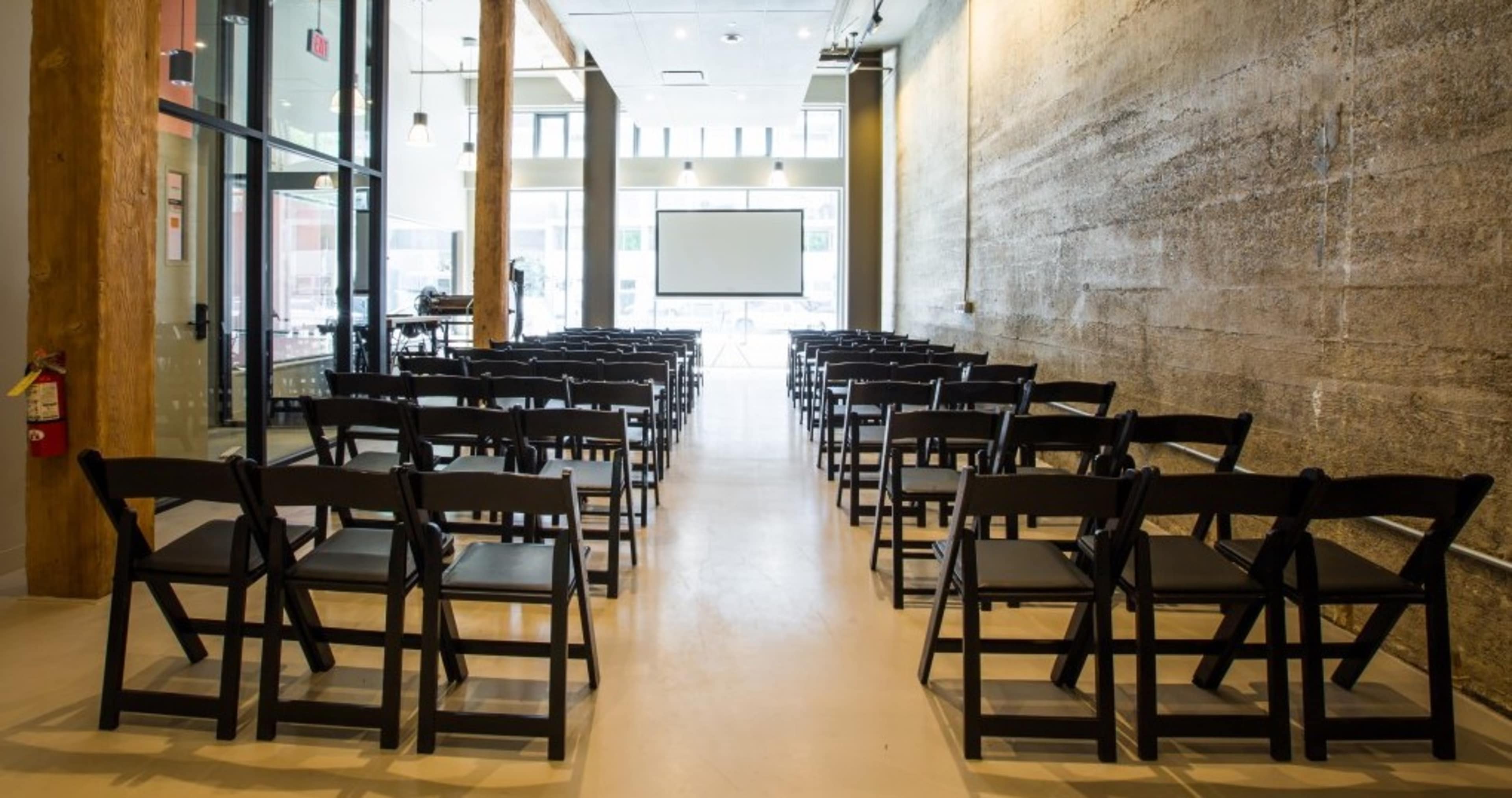 The image shows a conference room set up with rows of black folding chairs facing a projector screen against a concrete wall.
