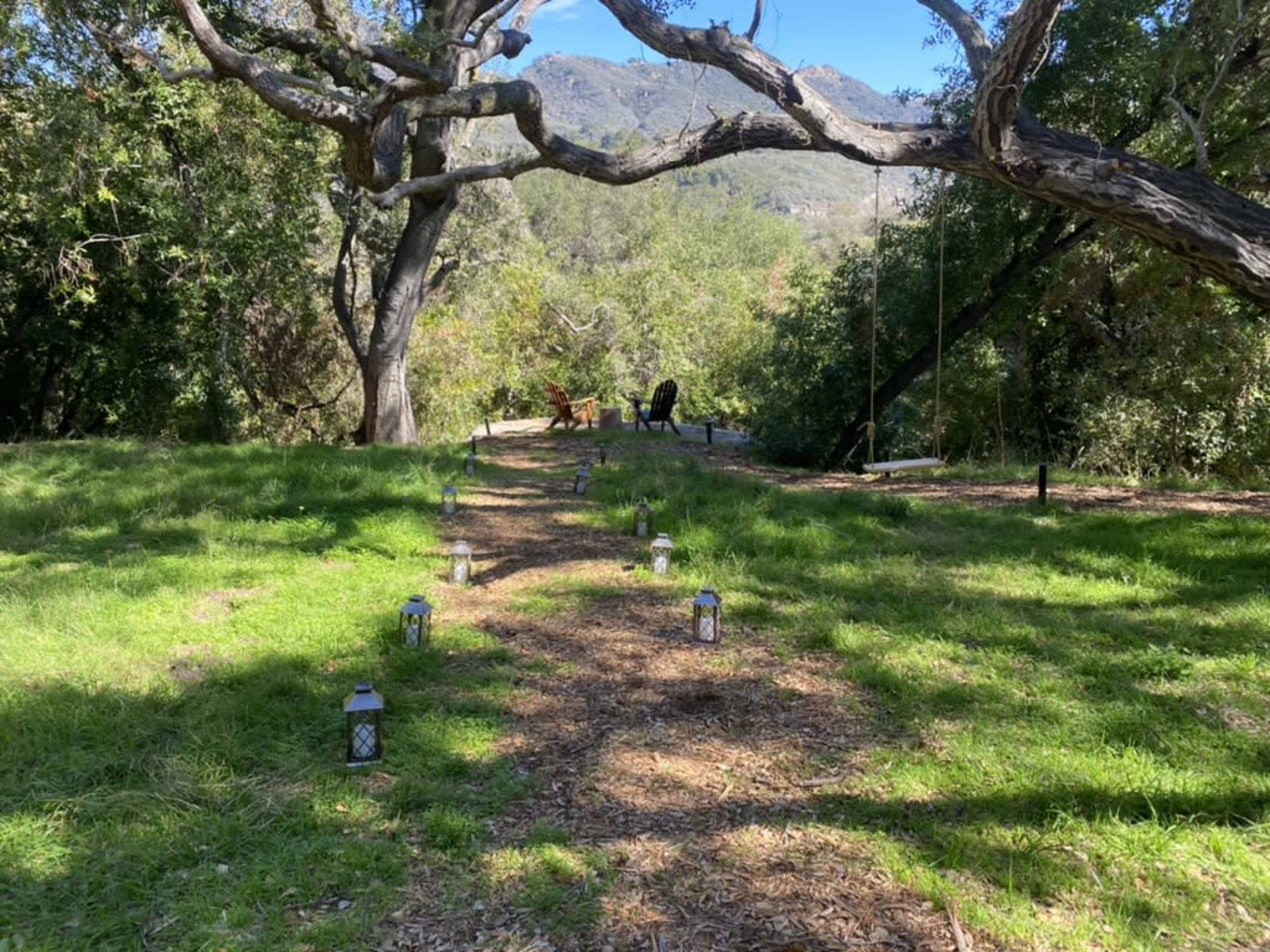 A pathway lined with lanterns leads to a seating area beneath large trees and mountains in the background.