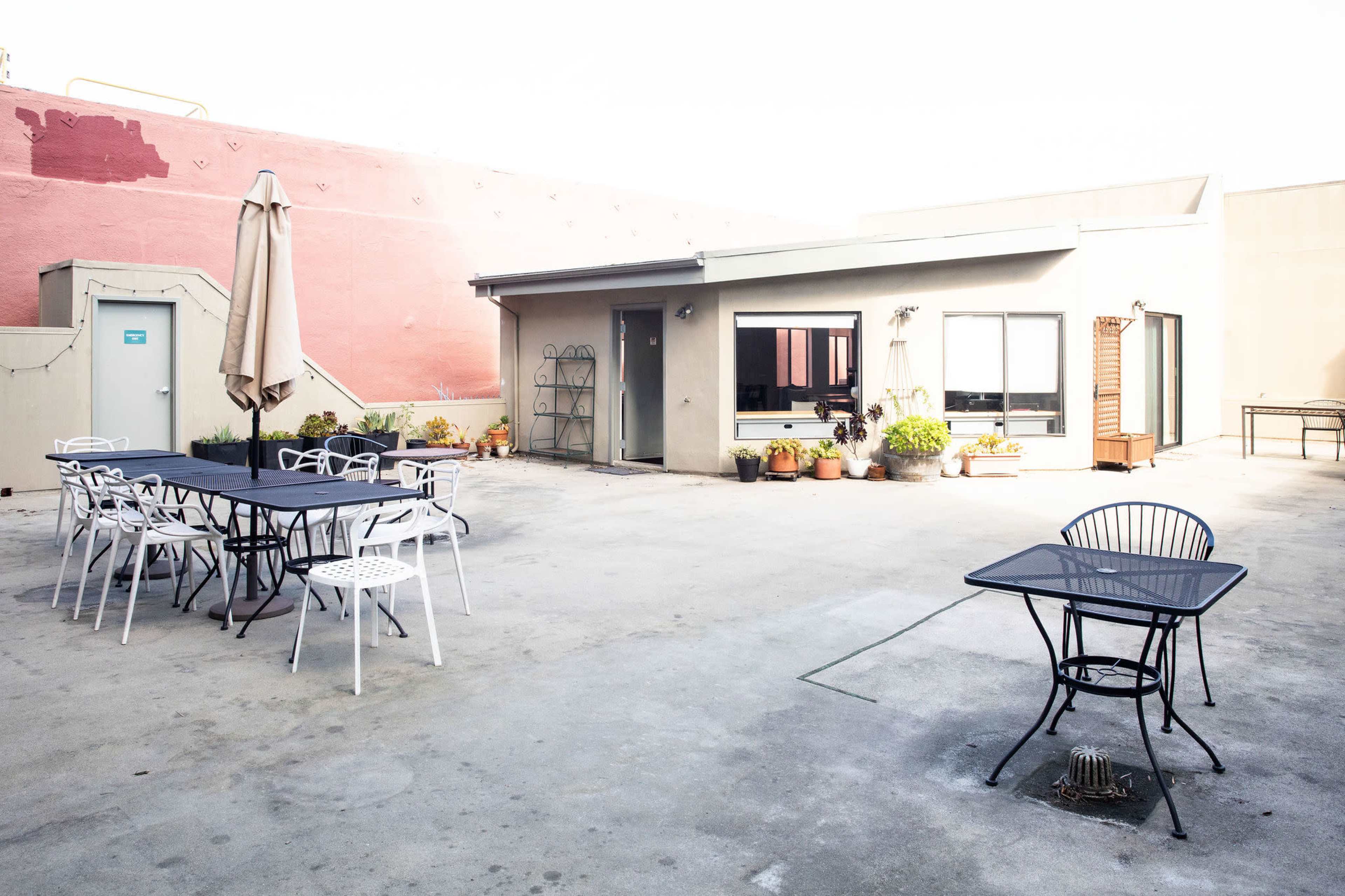 The image shows a rooftop patio with several tables and chairs, plants in pots, and a nearby building.