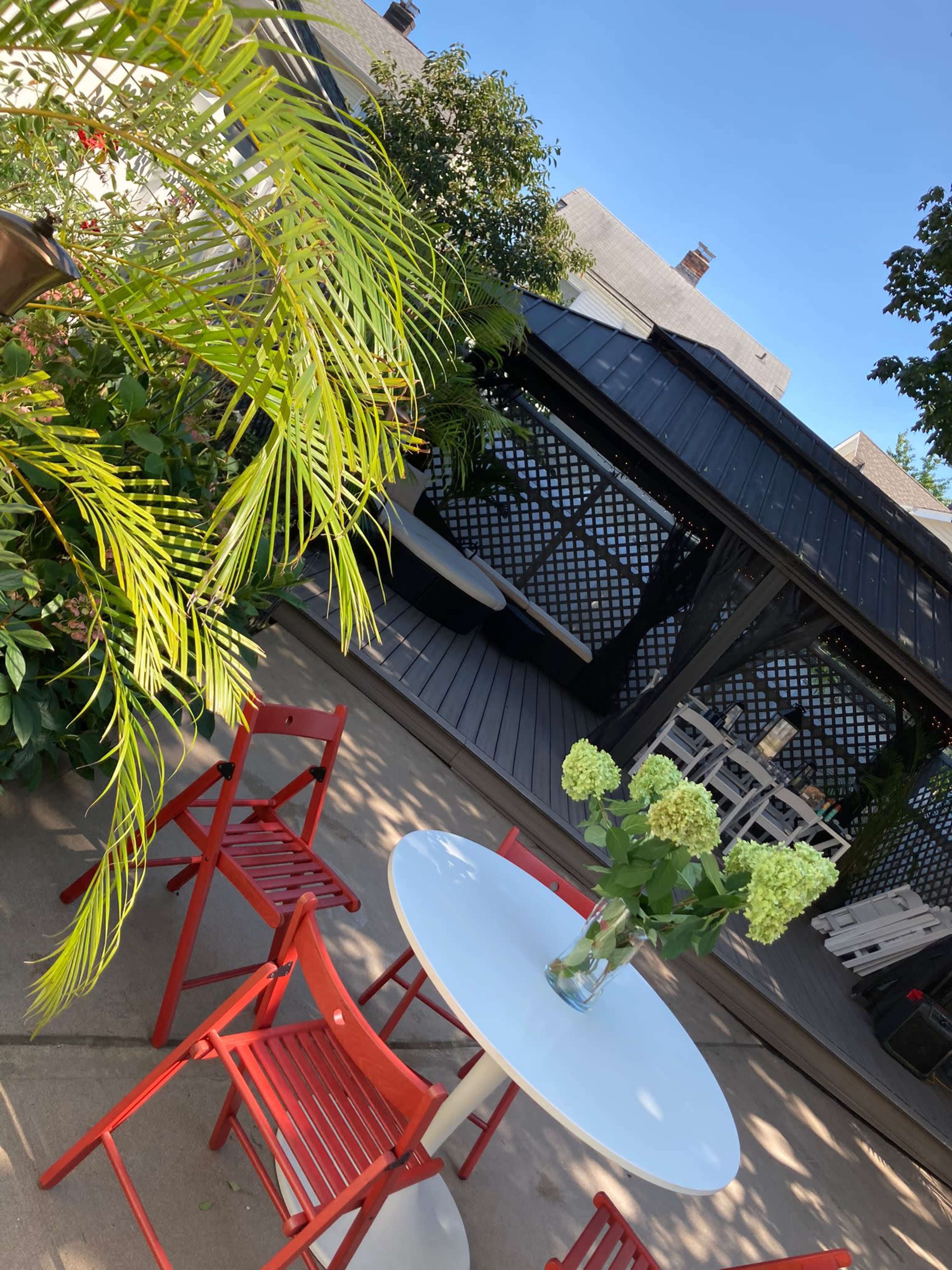 The image shows a patio area with a round white table and red chairs, surrounded by greenery and a shaded seating space in the background.