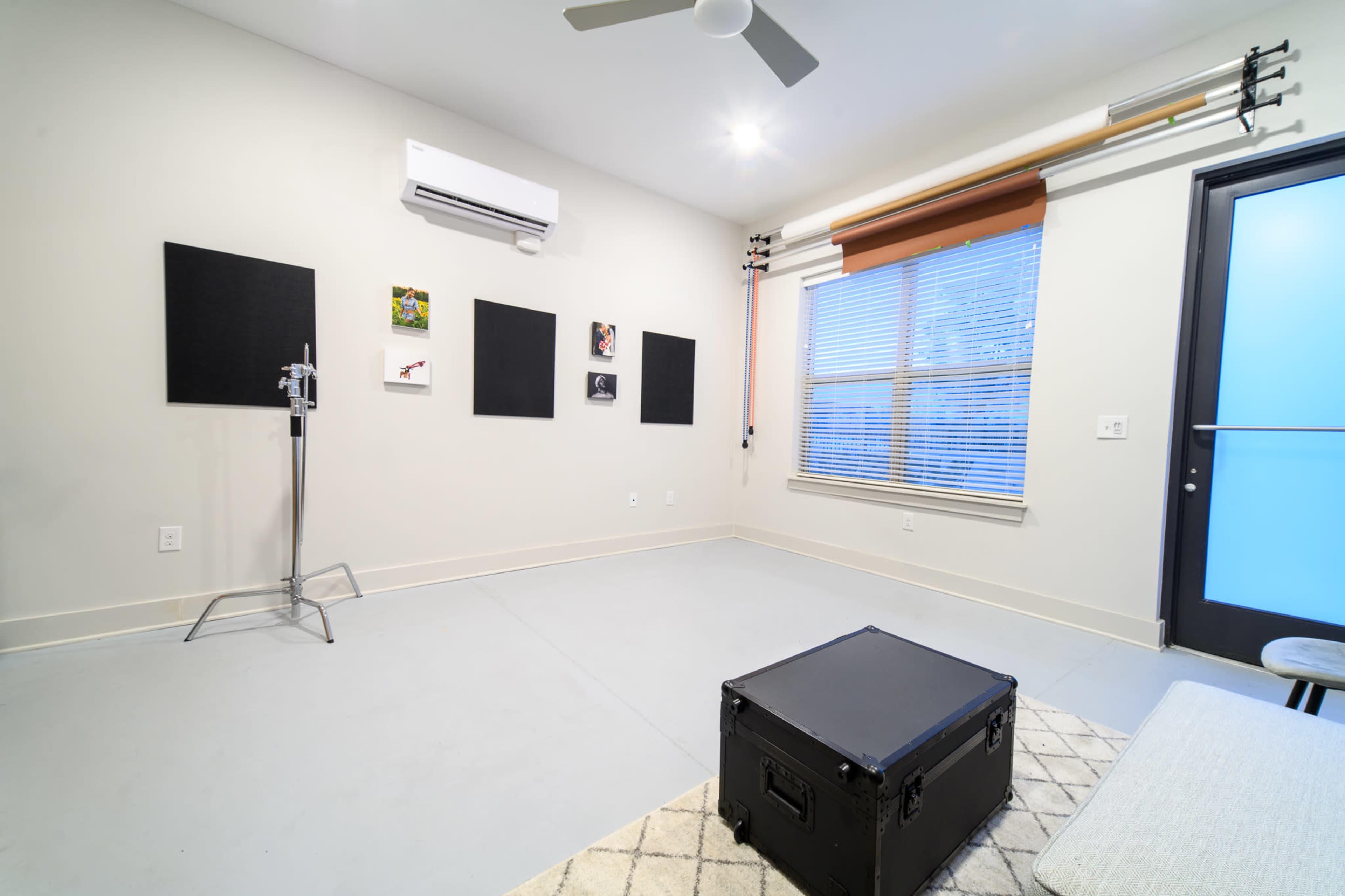 The image shows a sparsely furnished room with a concrete floor, featuring a black storage box, wall-mounted artwork, and a standing clothing rack.