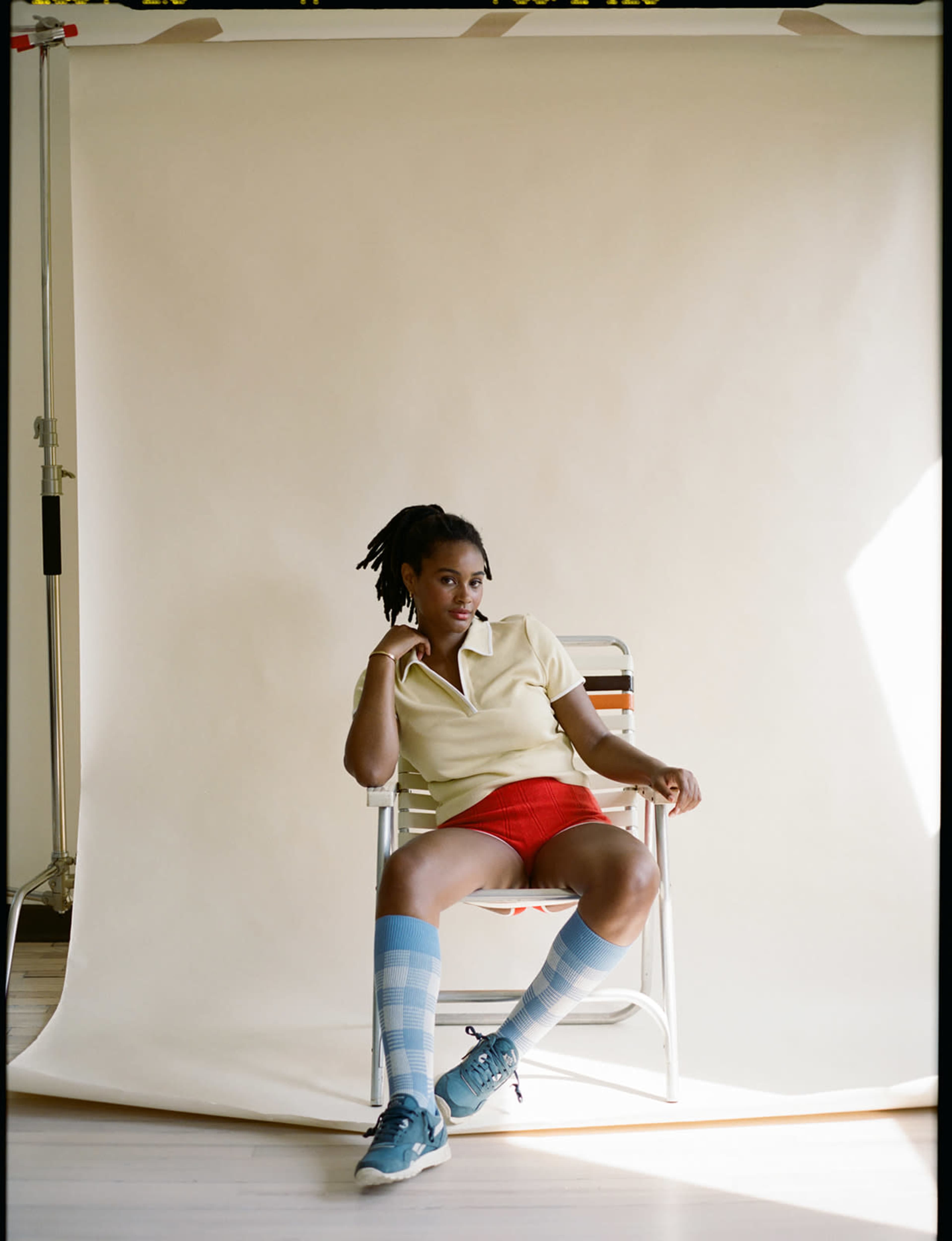 A person sits in a folding chair against a plain background, wearing a light yellow polo shirt, red shorts, and blue and white striped socks.
