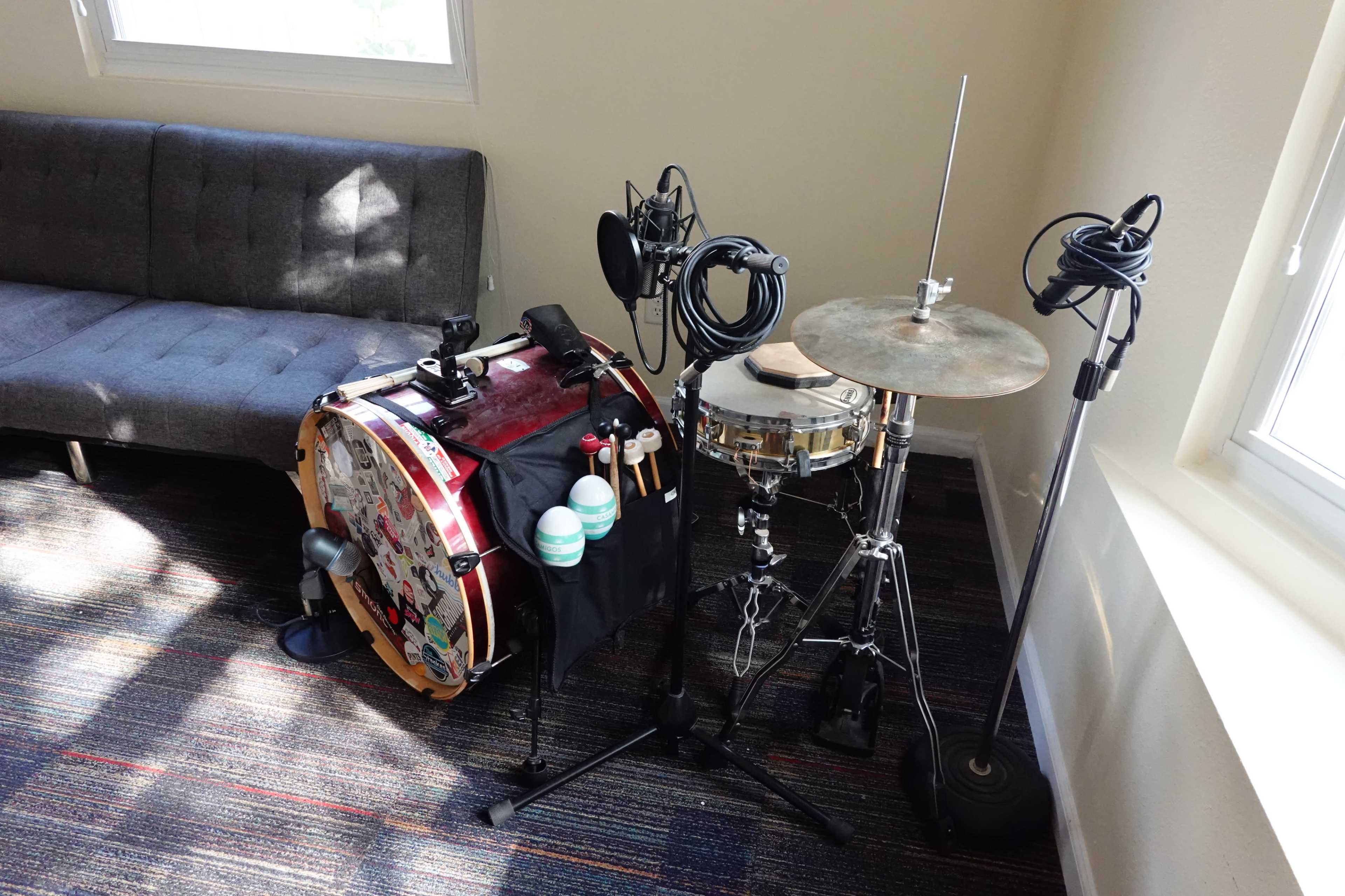 A drum set is set up next to a gray sofa in a well-lit room.