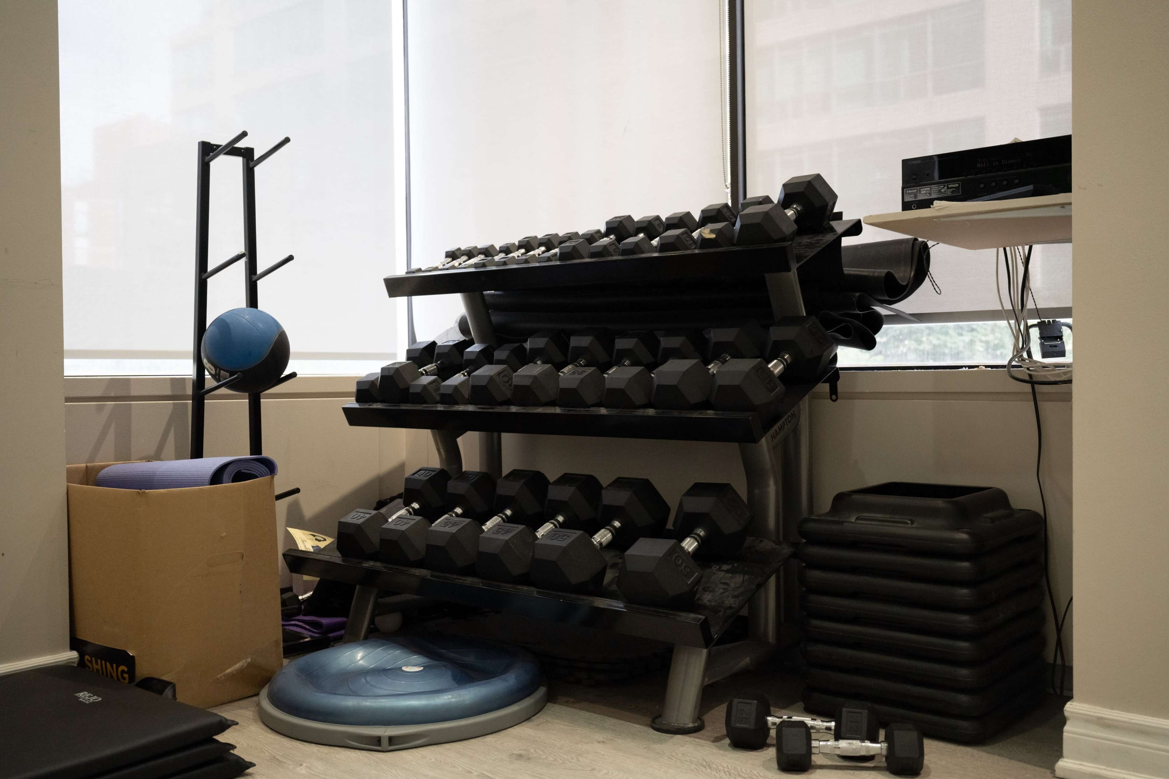 The image shows a small home gym area featuring a rack of dumbbells, exercise mats, a stability ball, and a stack of fitness steps.
