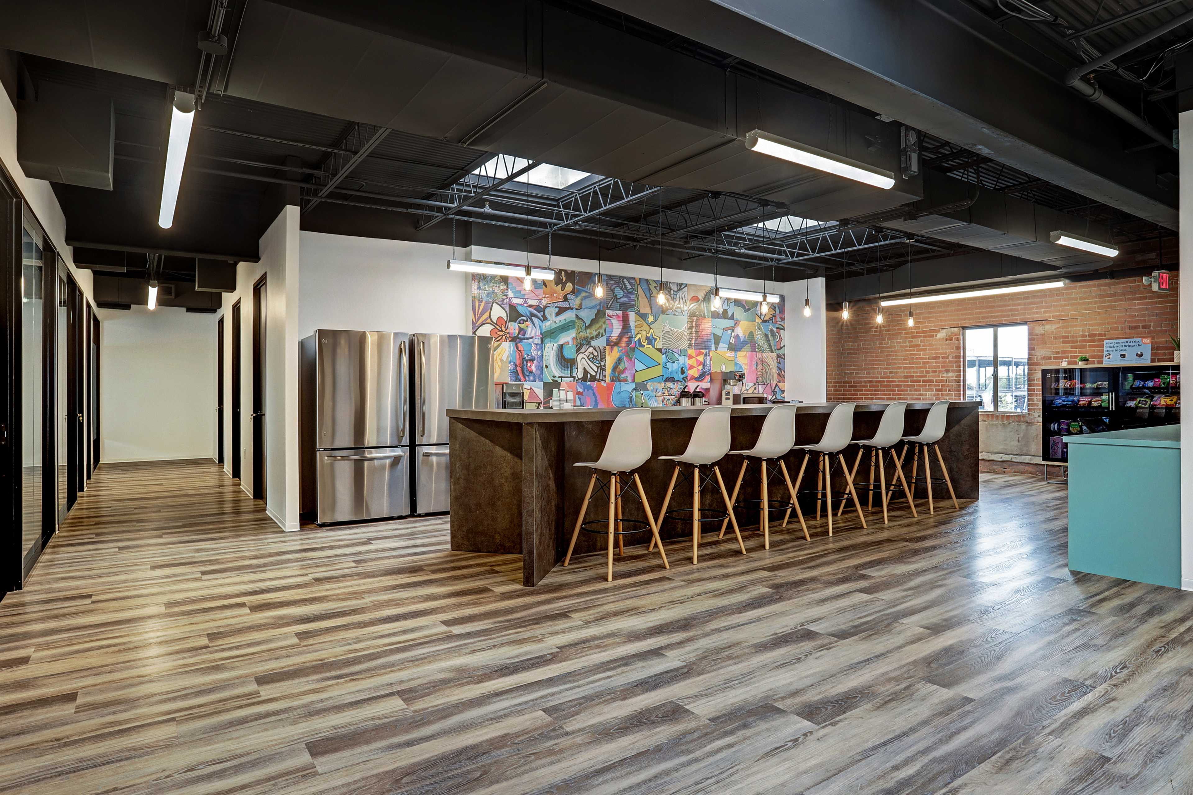 A modern office break area with a large bar counter, high stools, a colorful mural on the wall, and kitchen appliances in the background.