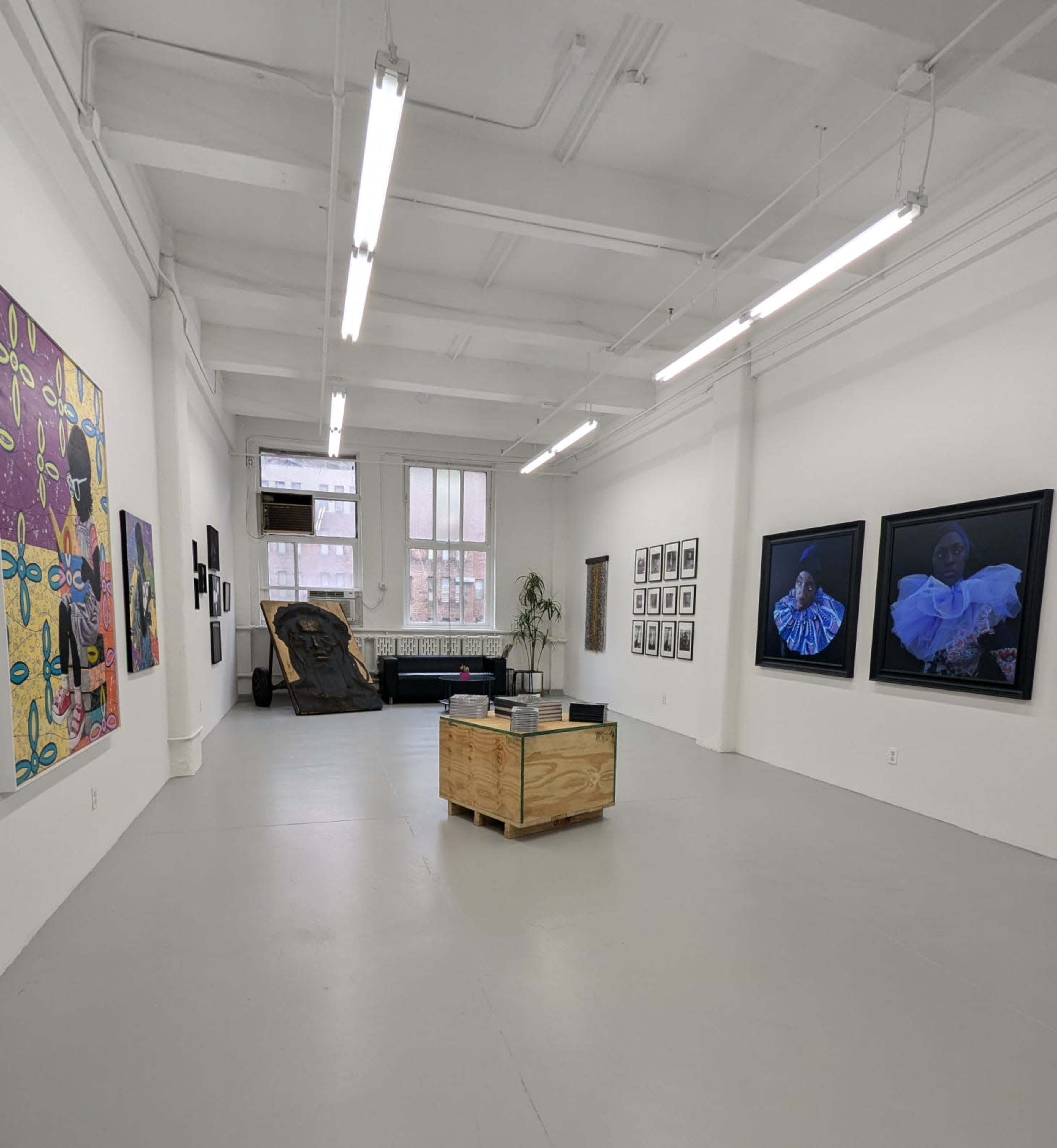 The image shows an art gallery with white walls, featuring various framed artworks, a wooden display table in the center, and a sculpture on one side.