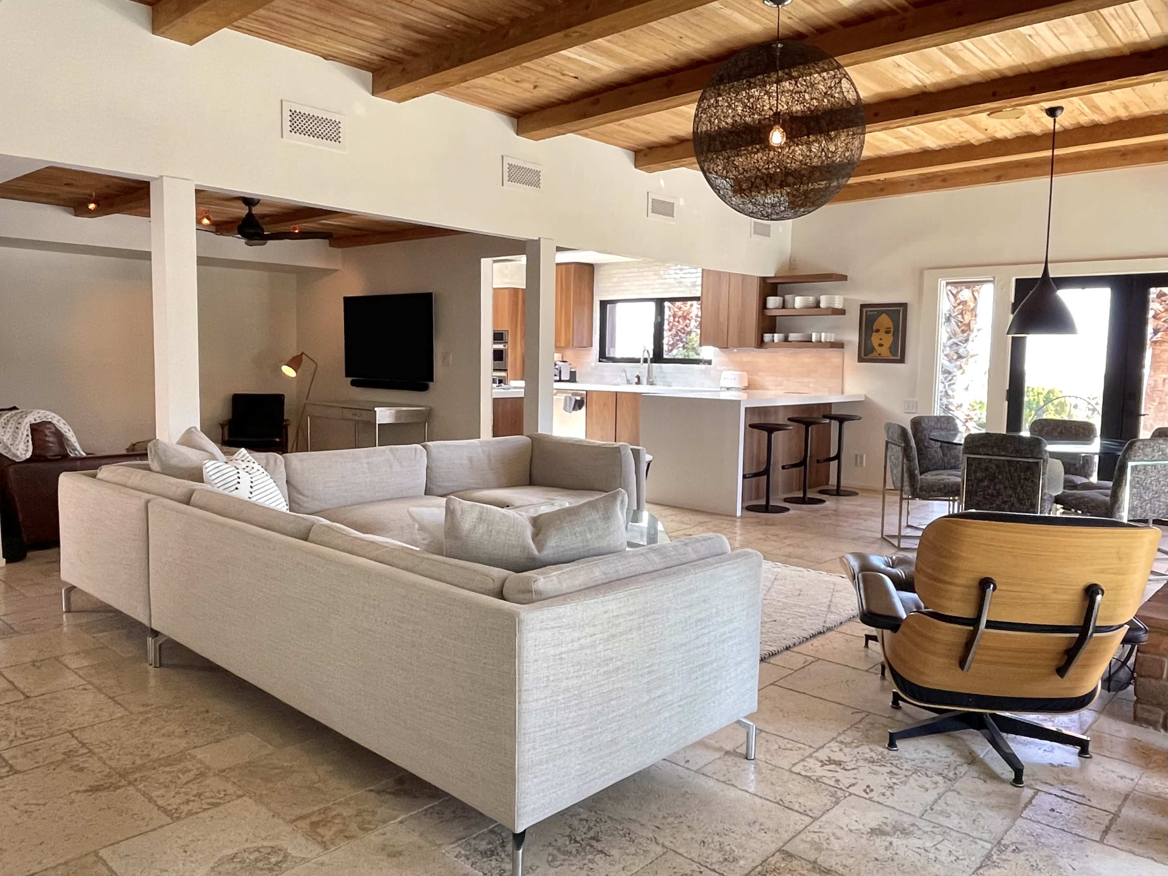 A modern living space featuring a white sectional sofa, wooden ceiling beams, and a dining area with a round table and chairs adjacent to a kitchen.