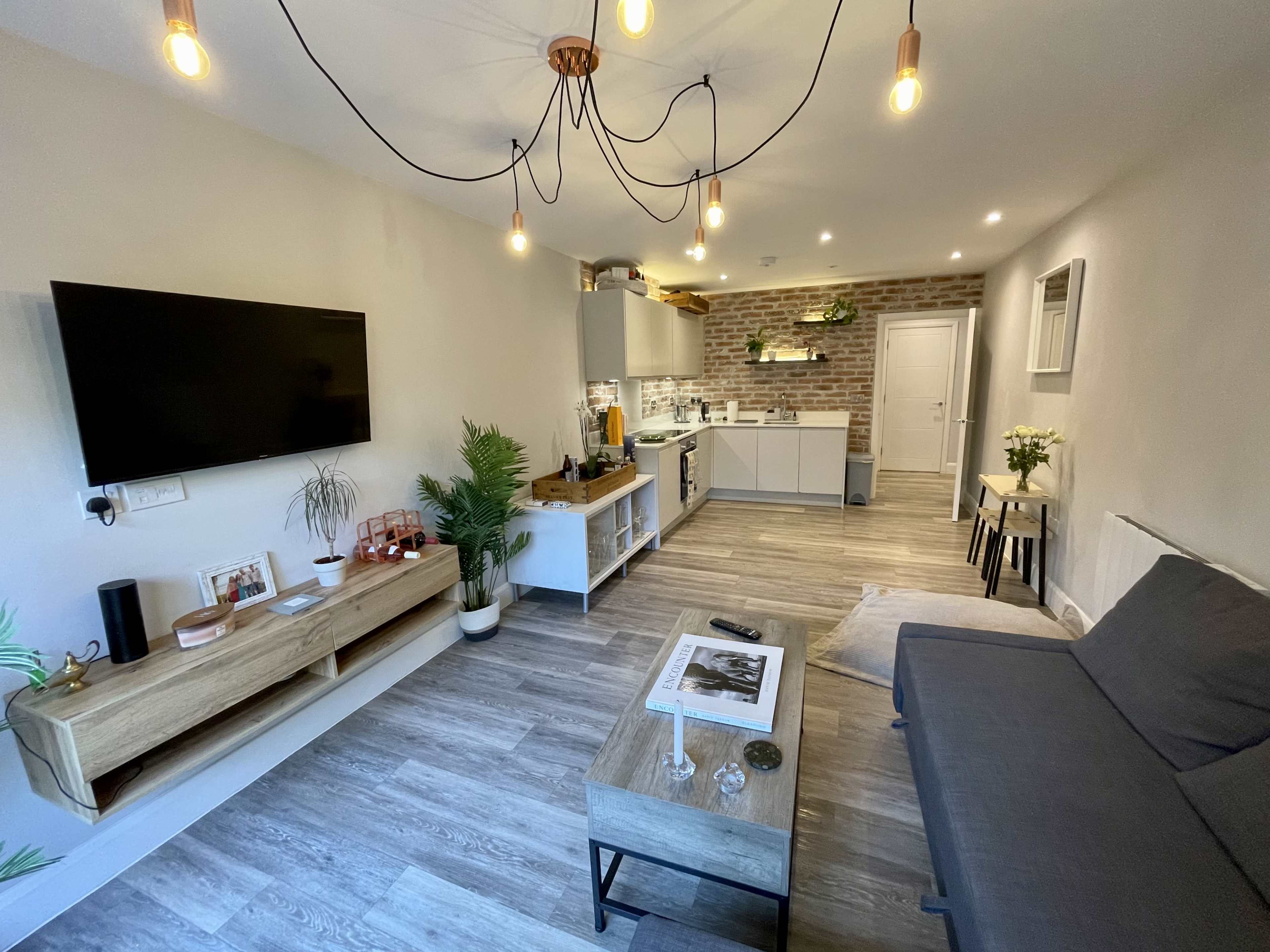 A modern living room with a gray sofa, a wooden coffee table, a TV mounted on the wall, and a kitchen area with brick accents visible in the background.