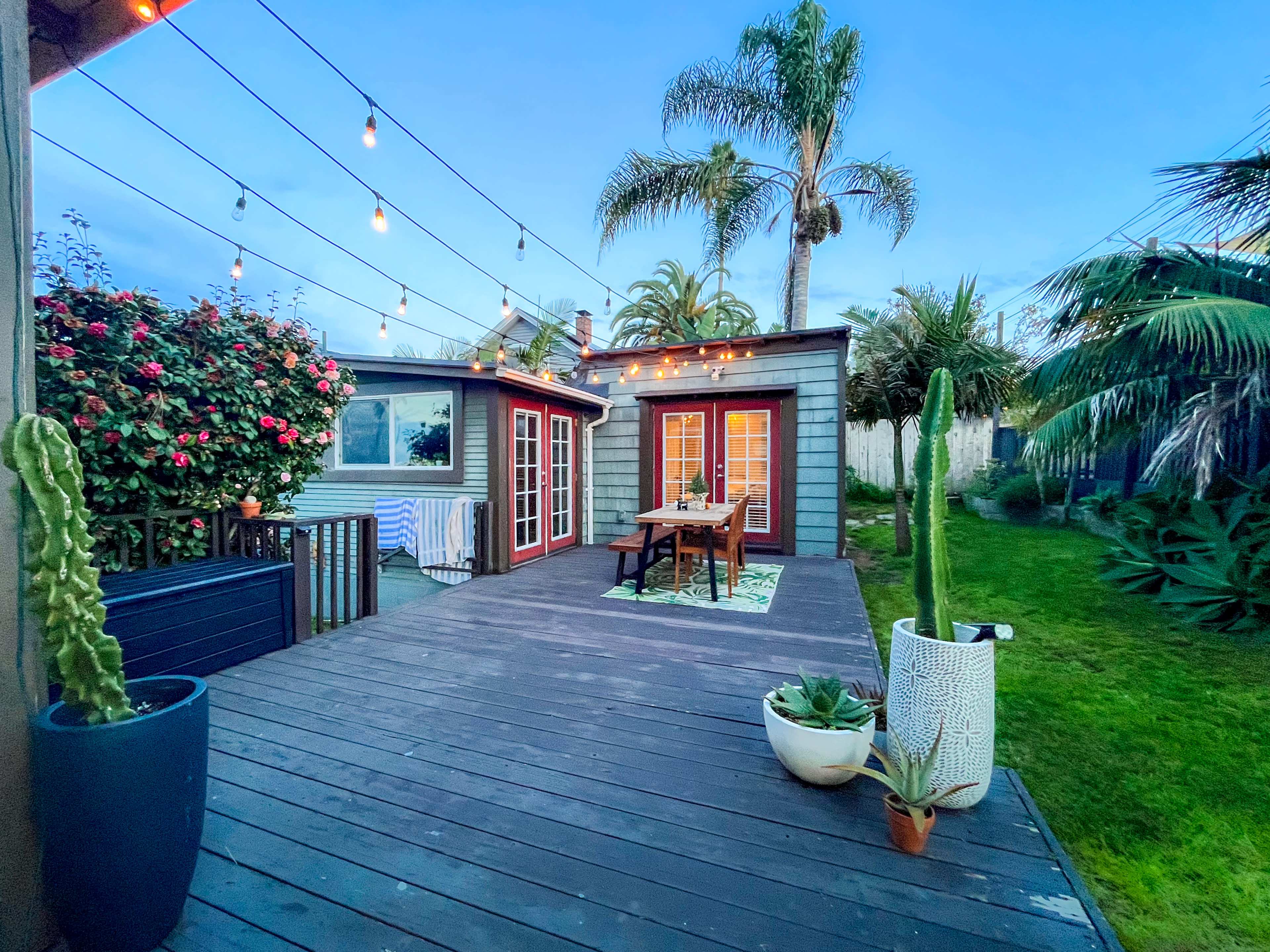 A wooden deck features a dining table surrounded by decorative plants, with a cozy cabin and string lights overhead in a garden setting.