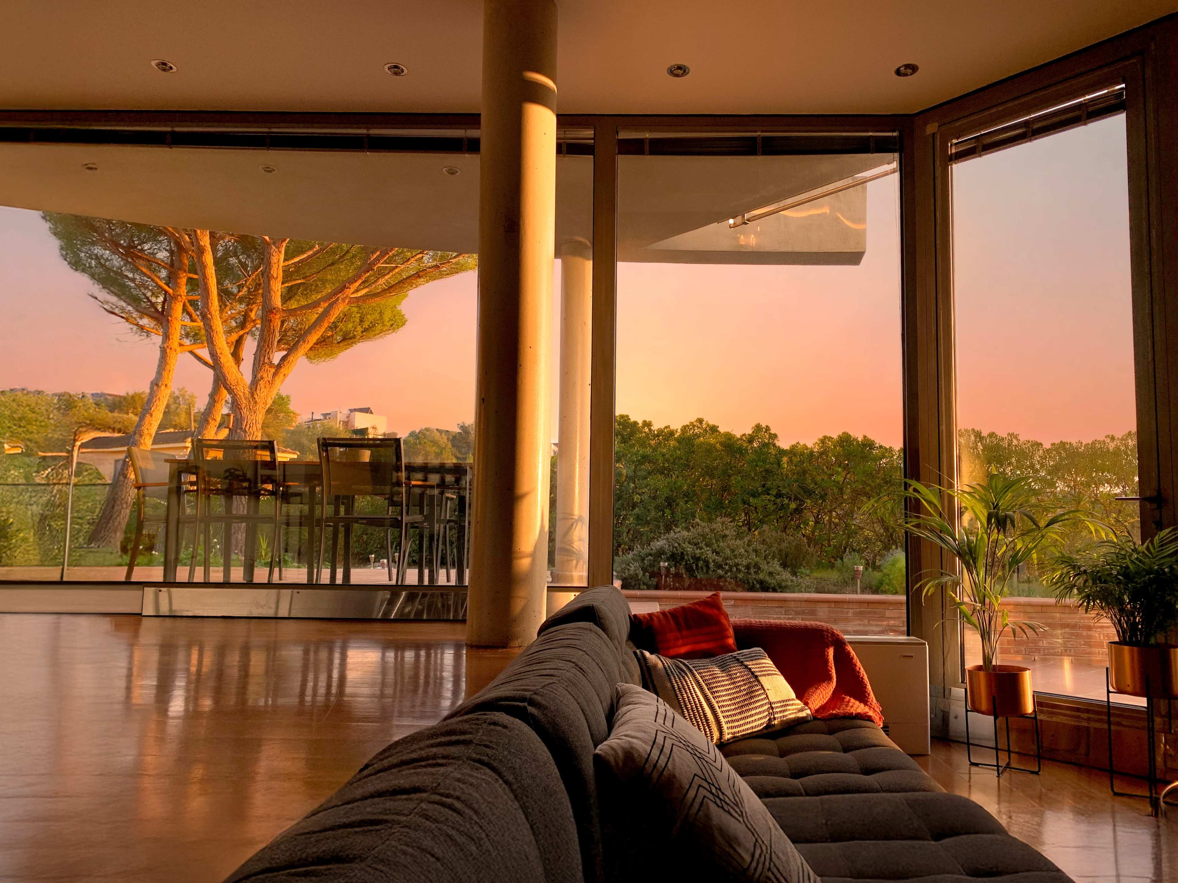 The image shows a modern living room with a large window overlooking a garden at sunset, featuring a sofa and a dining area in the background.