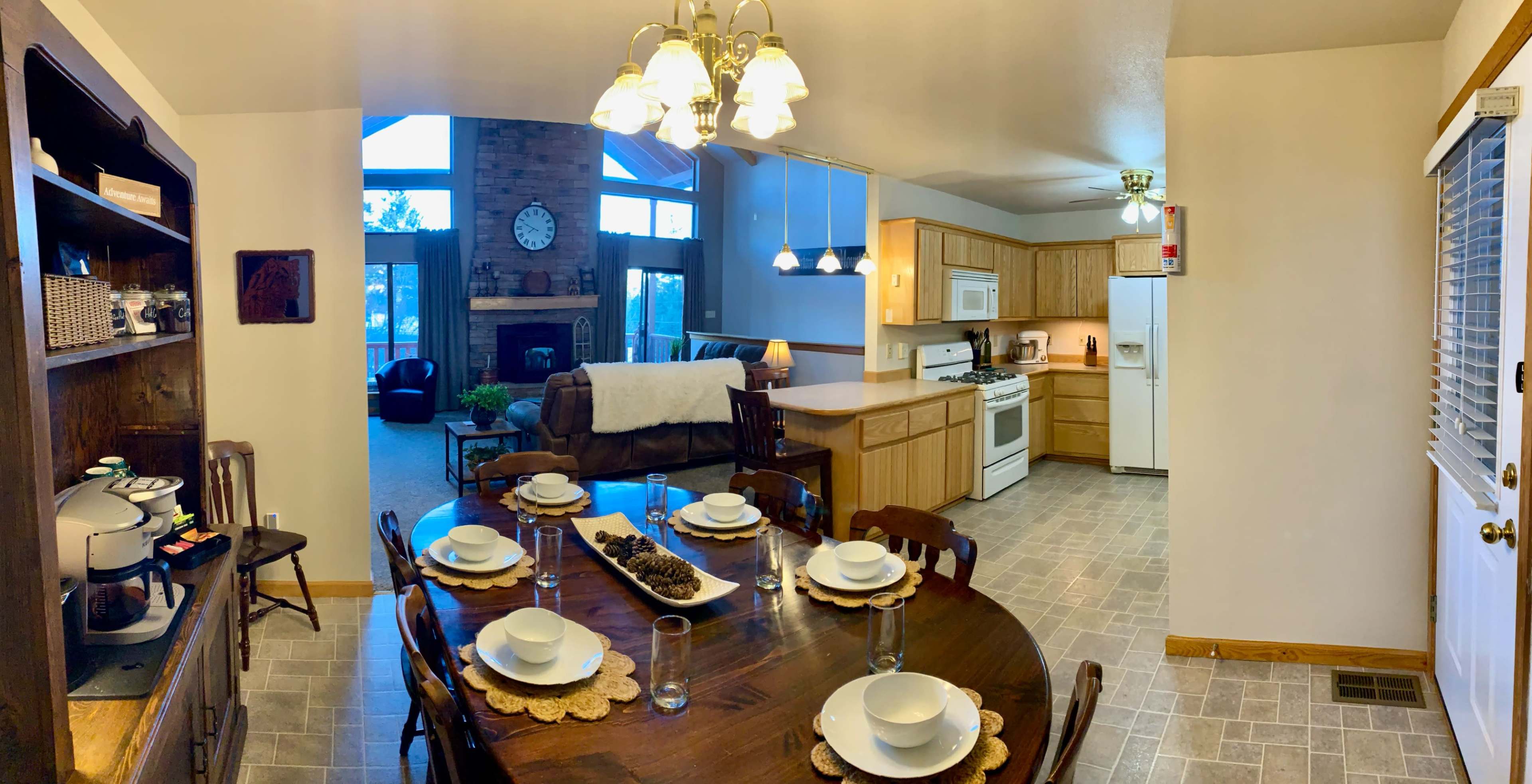 The image shows a spacious kitchen and dining area with a large wooden table set for a meal, adjacent to a living room featuring a brick fireplace and large windows.