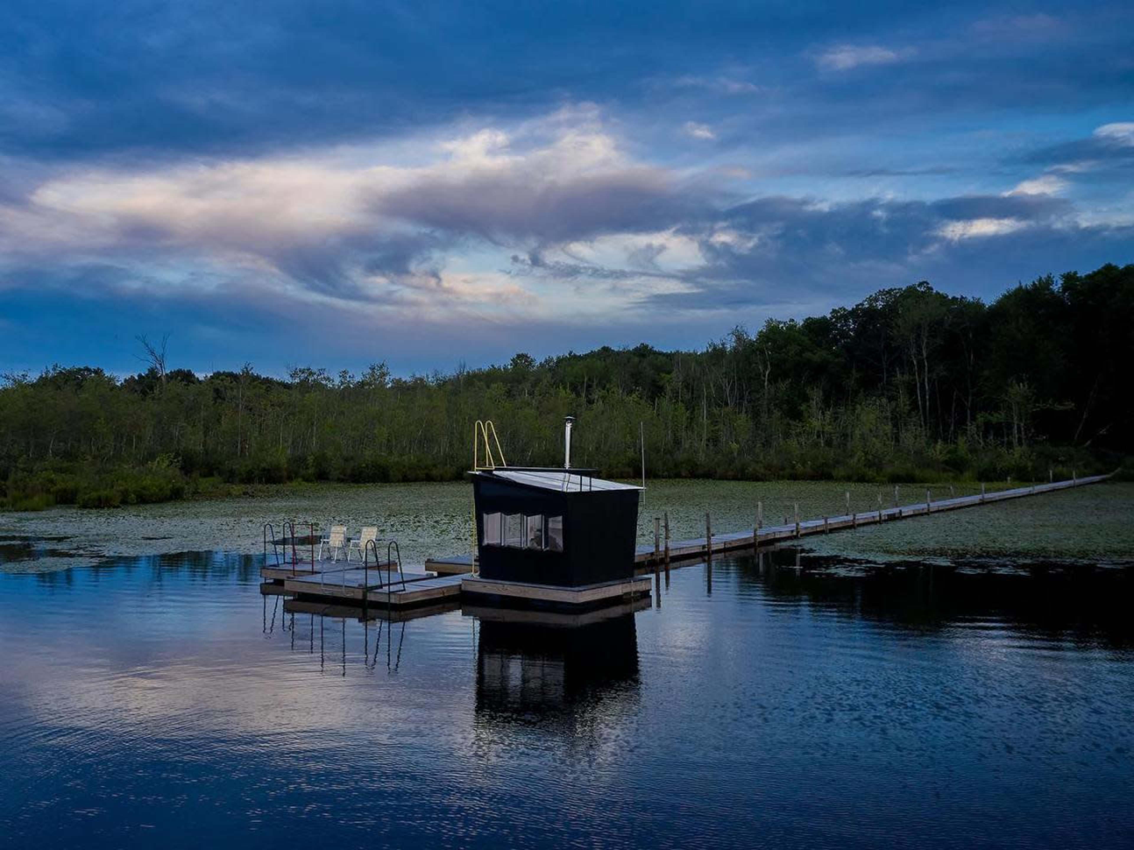 A dock extends into a calm body of water, featuring a small structure surrounded by greenery and a cloudy sky.