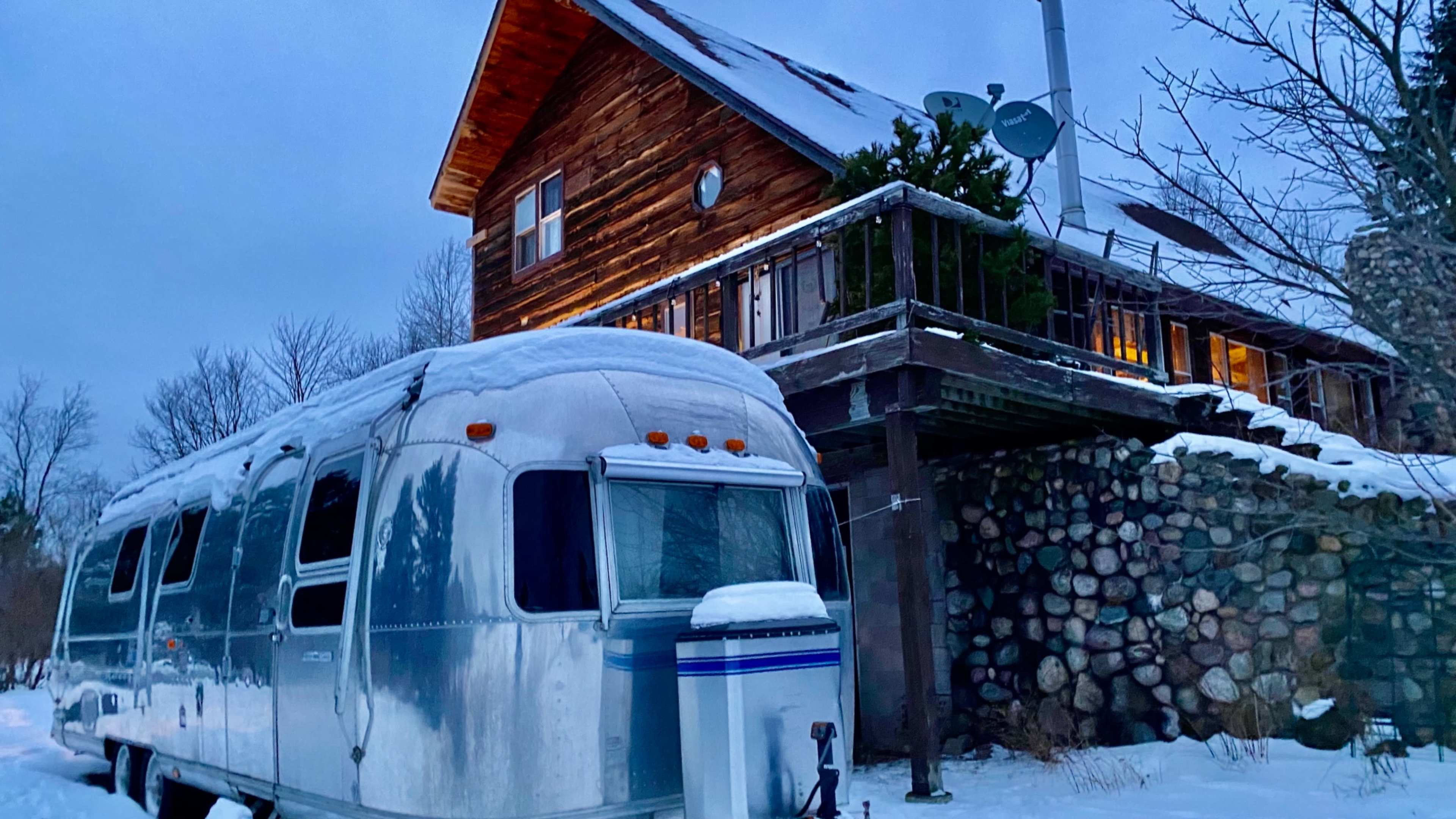 An Airstream trailer parked in a snow-covered landscape next to a wooden house with a balcony and satellite dishes.