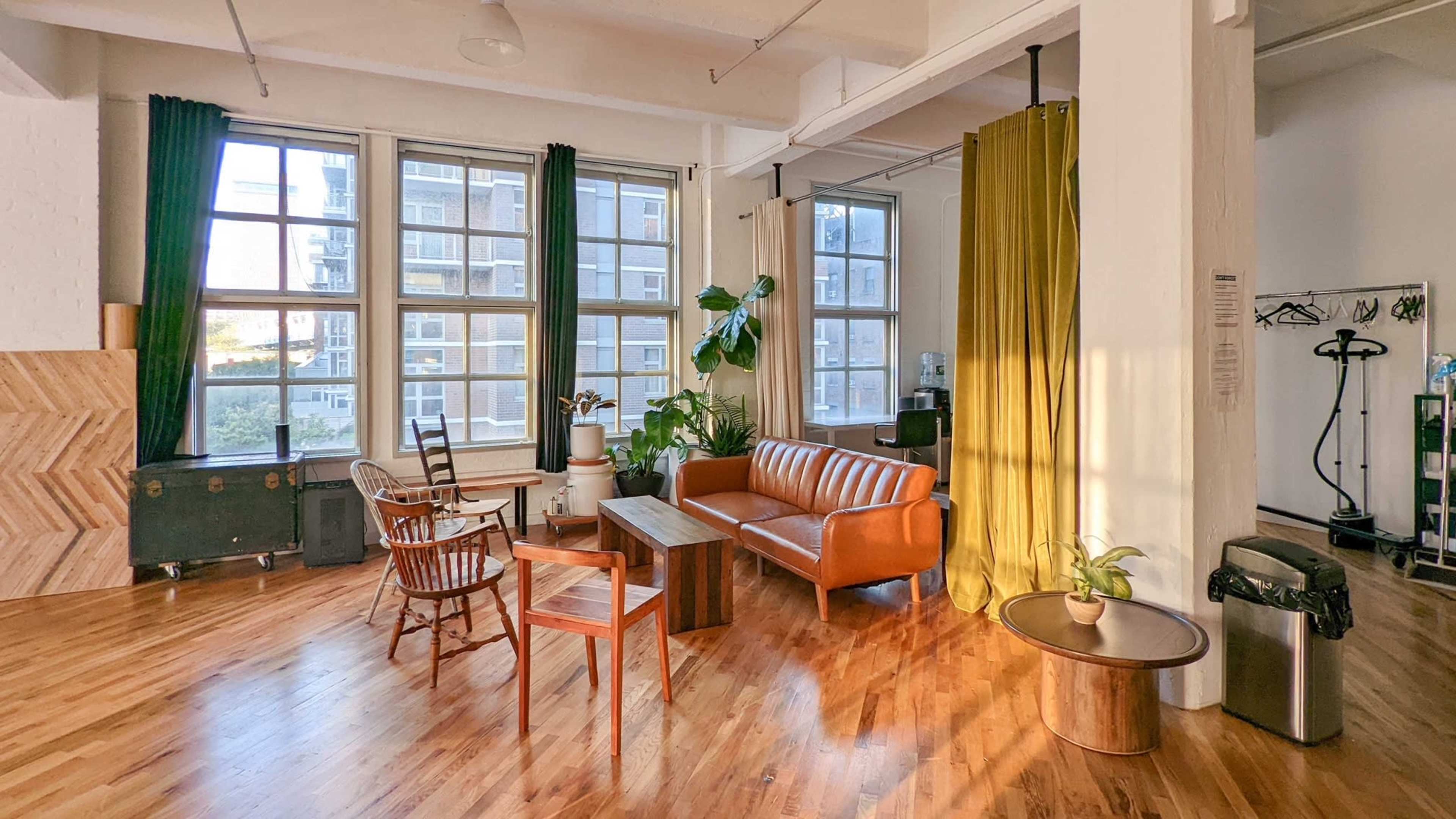 A bright, spacious room with large windows, featuring a brown leather sofa, wooden coffee table, and plants, along with a mix of green and gold curtains.