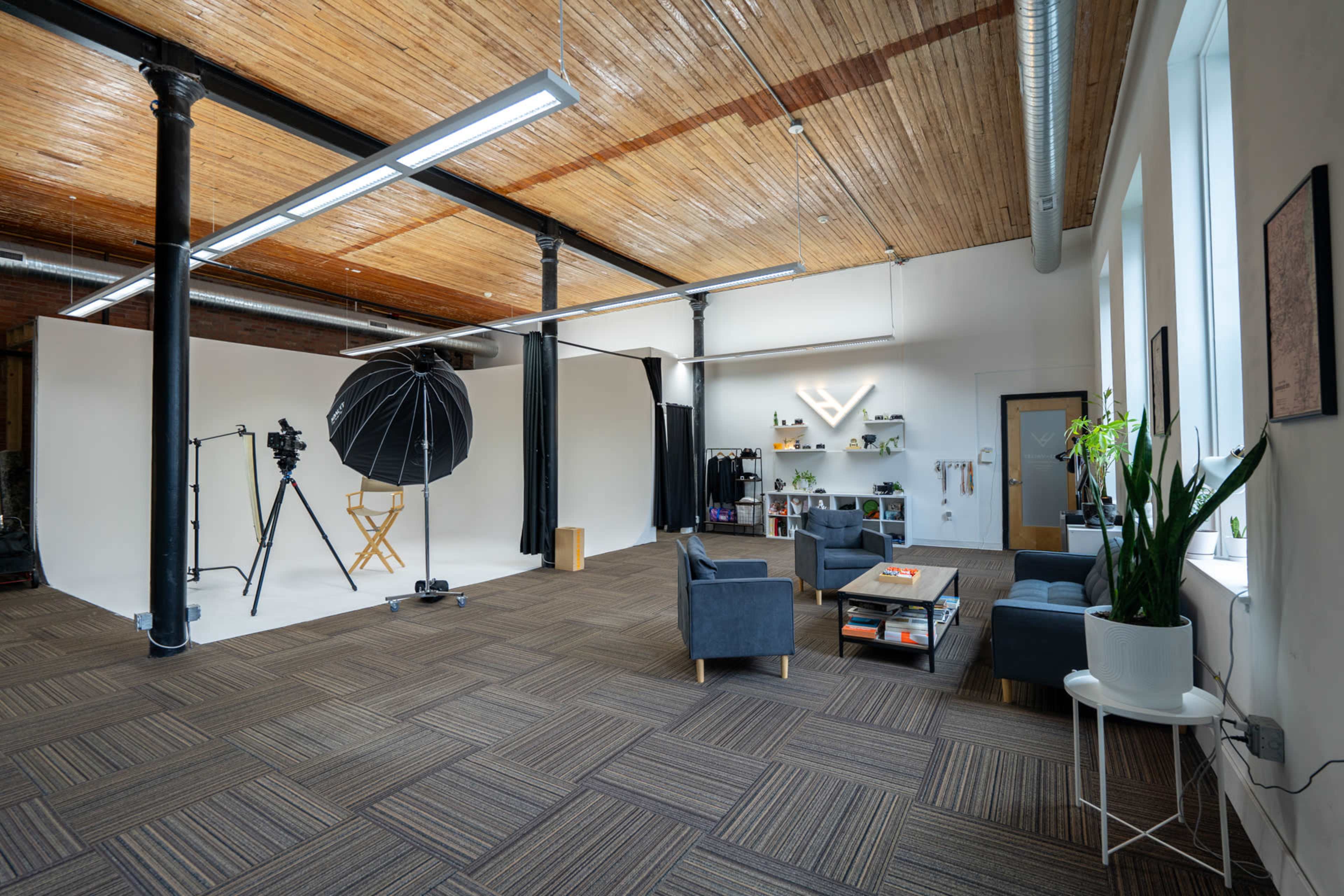 A spacious studio with a white backdrop, photography equipment, and a seating area furnished with gray sofas and decorative plants.