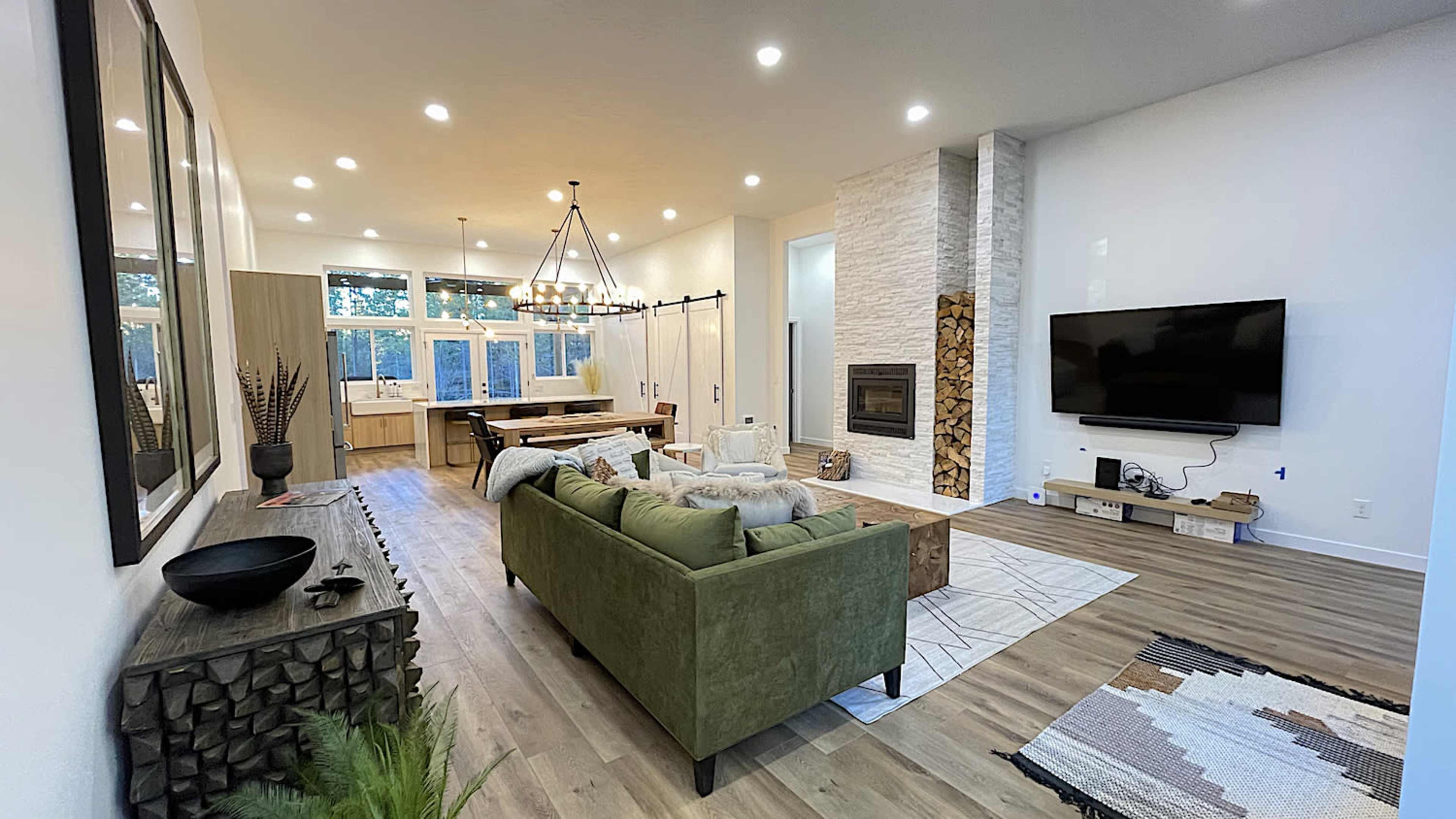A spacious living room features a green sofa, a wooden dining table, and a stone fireplace, with large windows letting in natural light.