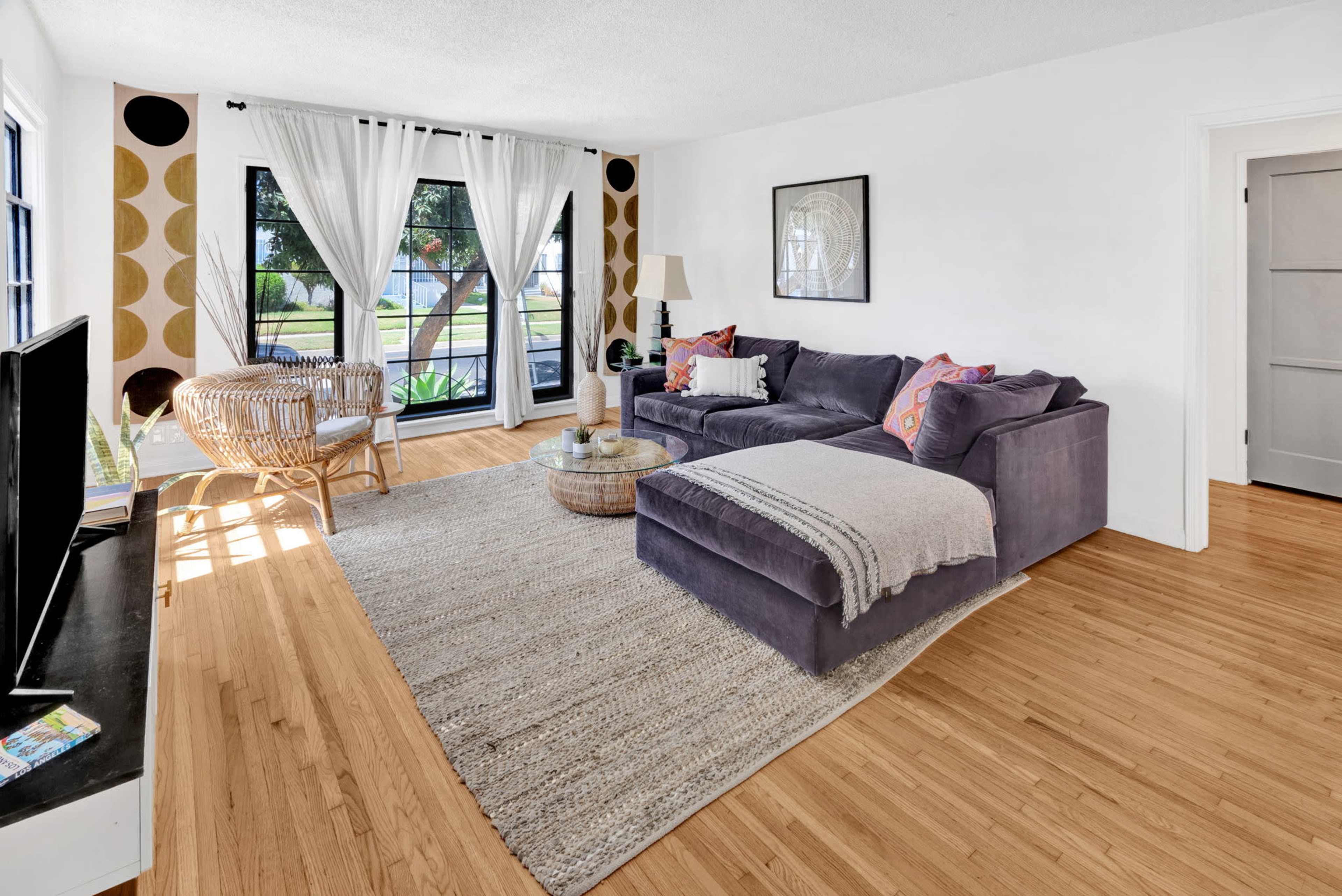 The living room features a grey sectional sofa, light hardwood flooring, large windows with sheer curtains, and a round coffee table in front of a television.