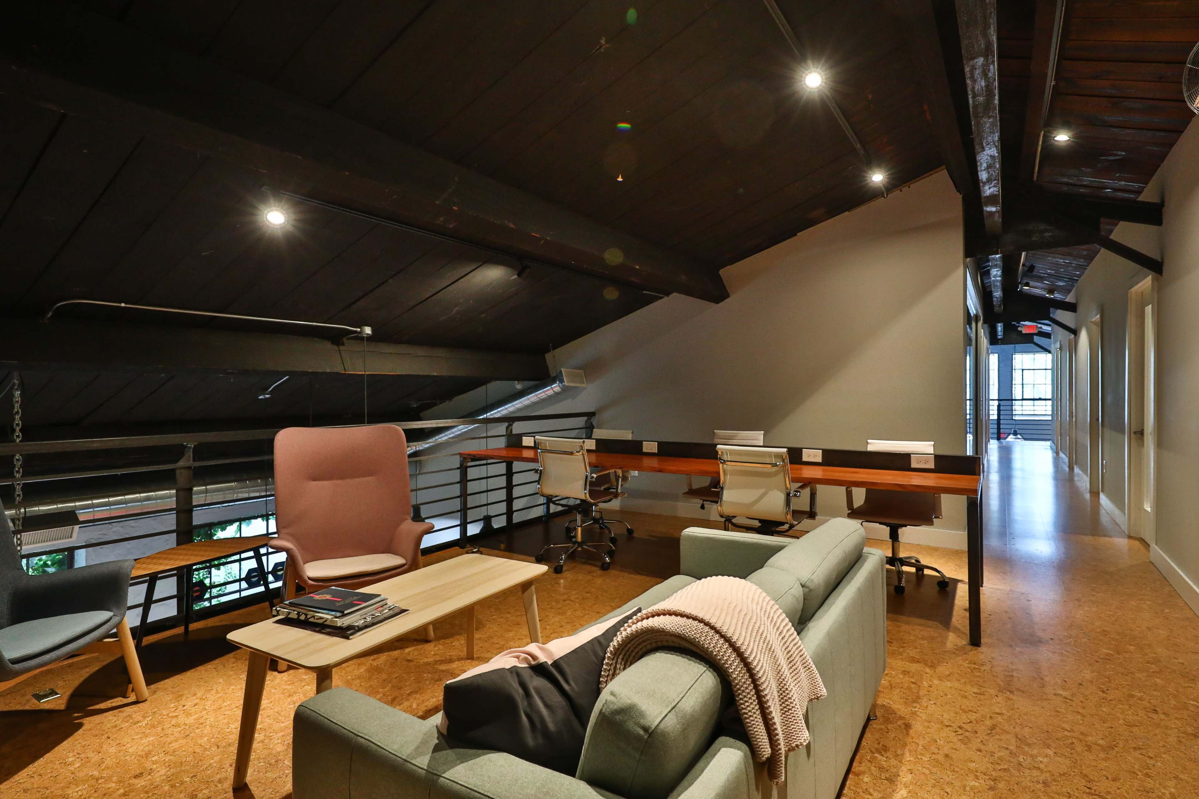 A modern loft-style workspace featuring a light-colored sofa, a coffee table with magazines, and a row of office chairs along a wooden tabletop.