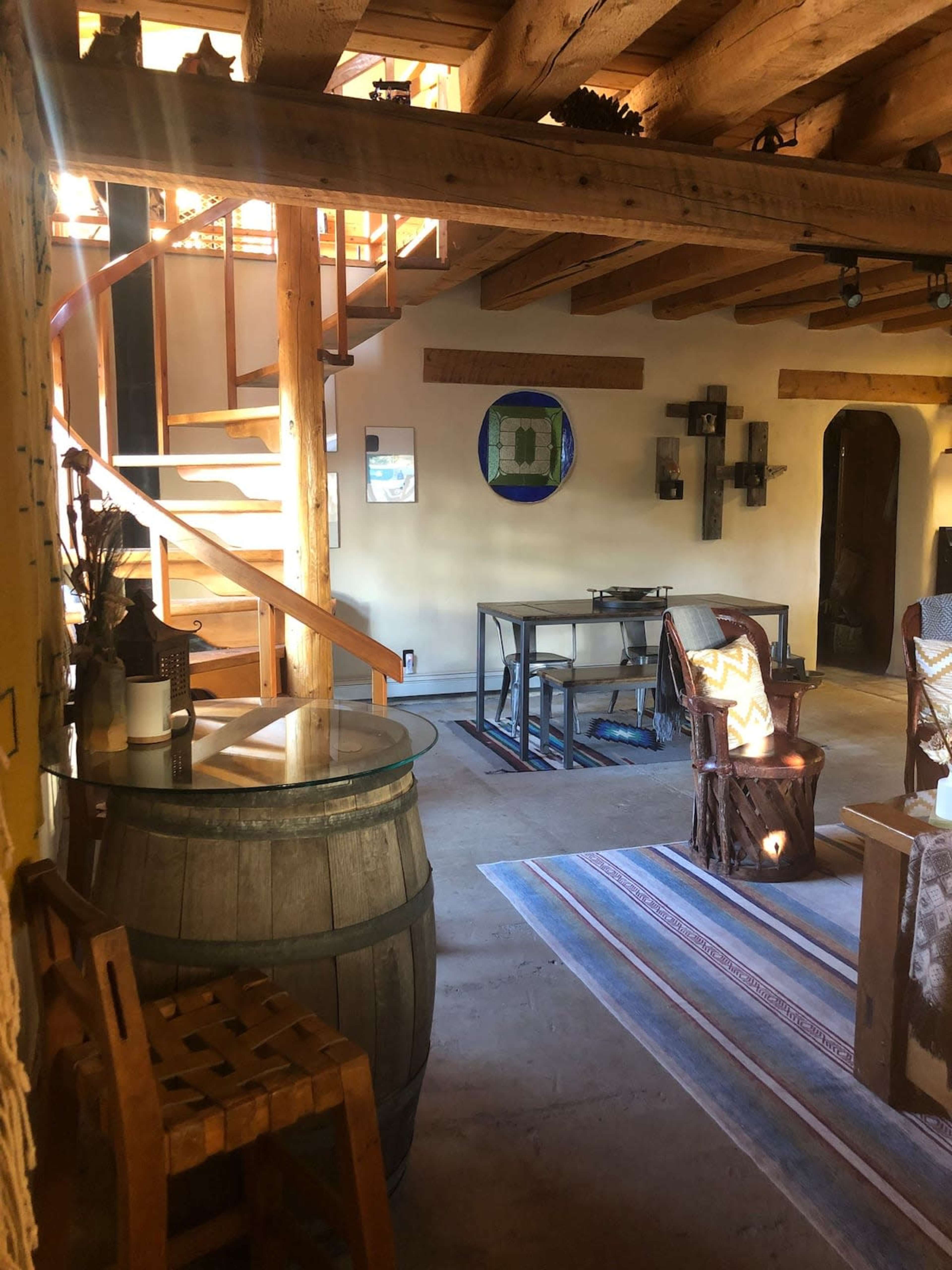 A rustic interior with exposed wooden beams, a wooden staircase, a dining table, and seating arranged around a patterned rug.
