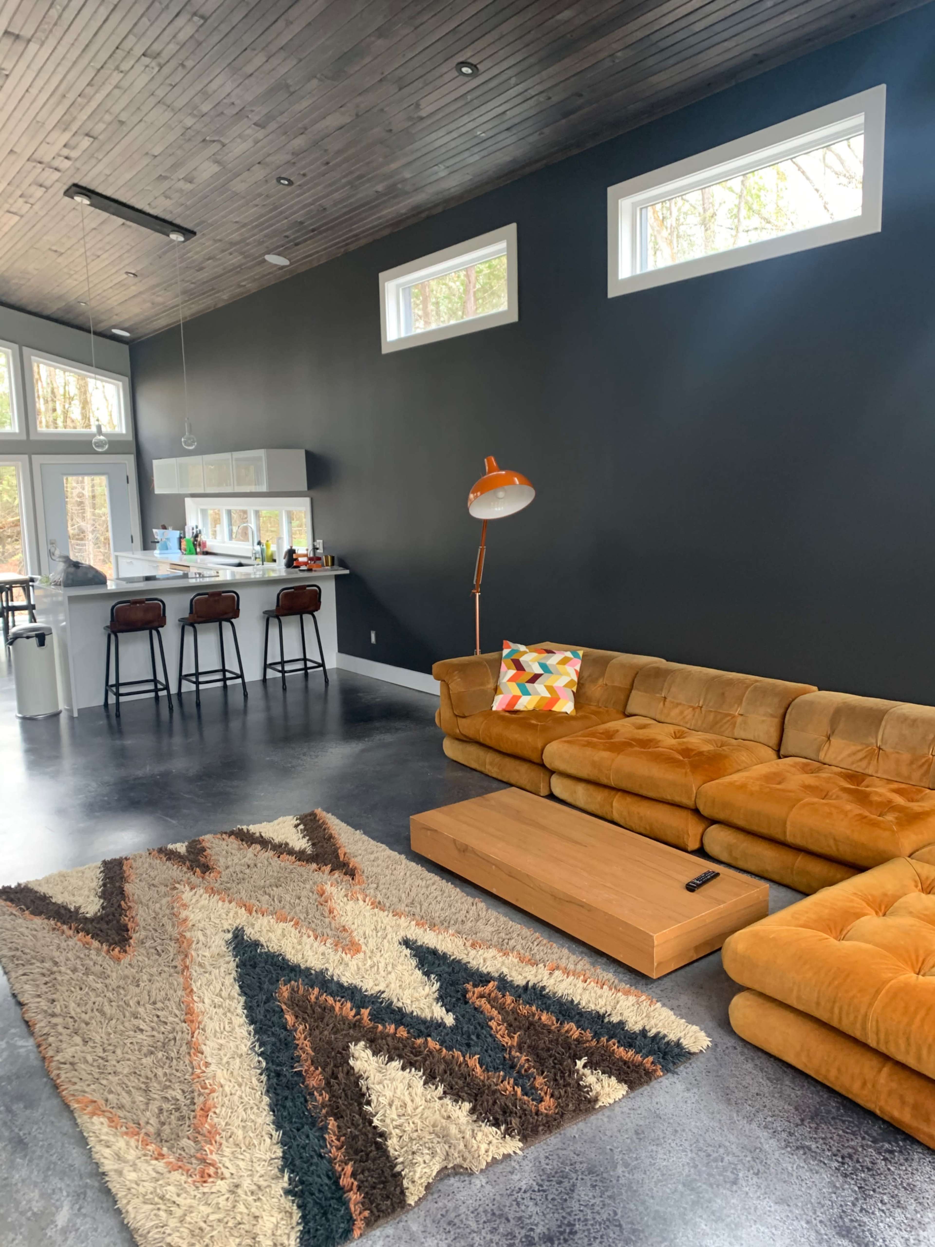 A modern living room with a brown sectional sofa, a wooden coffee table, and a patterned rug, along with a kitchen area visible in the background.