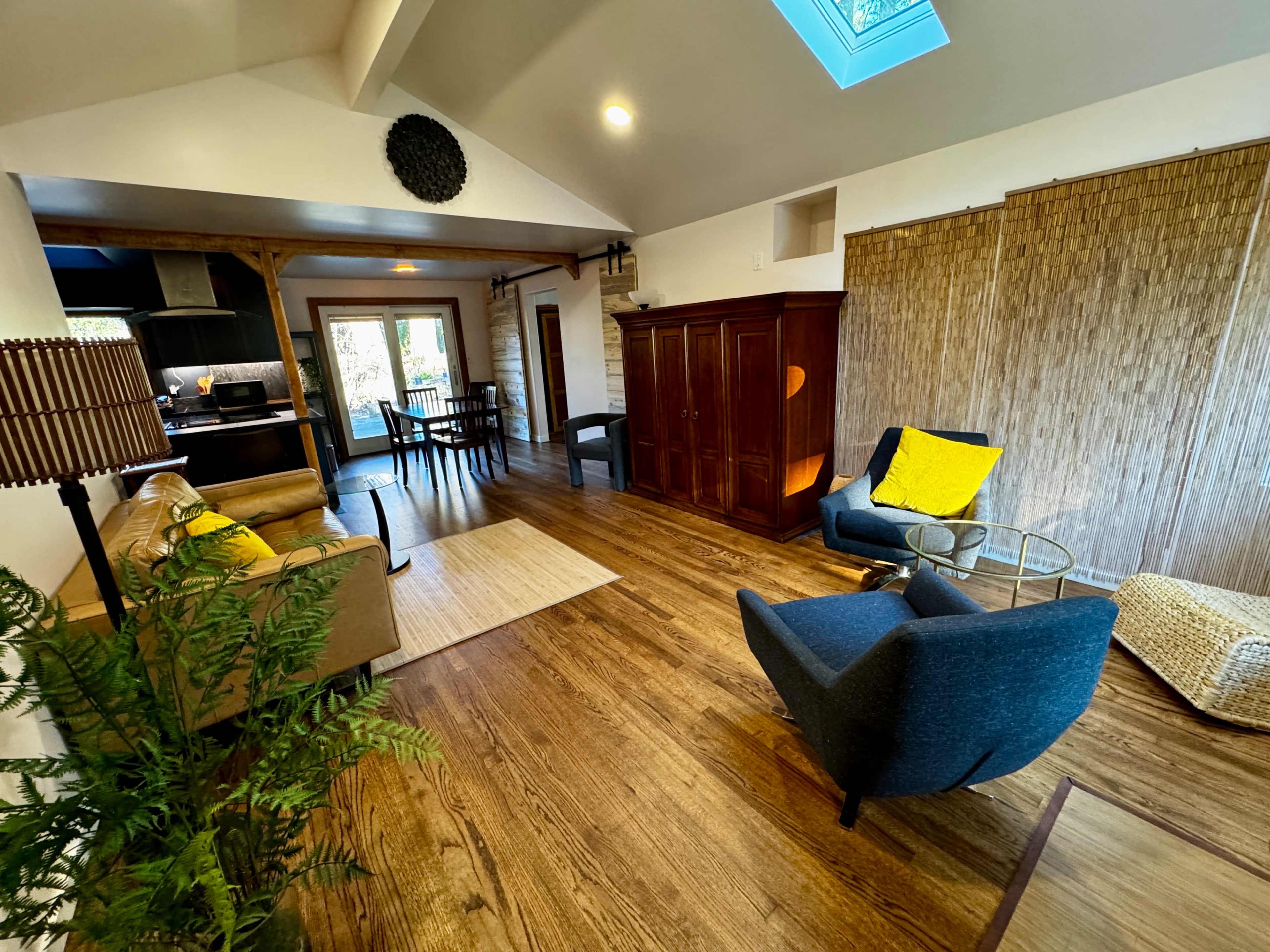 The image shows a spacious, well-lit living area featuring a couch, two chairs, a dining table, and wooden flooring with a decorative screen in the background.